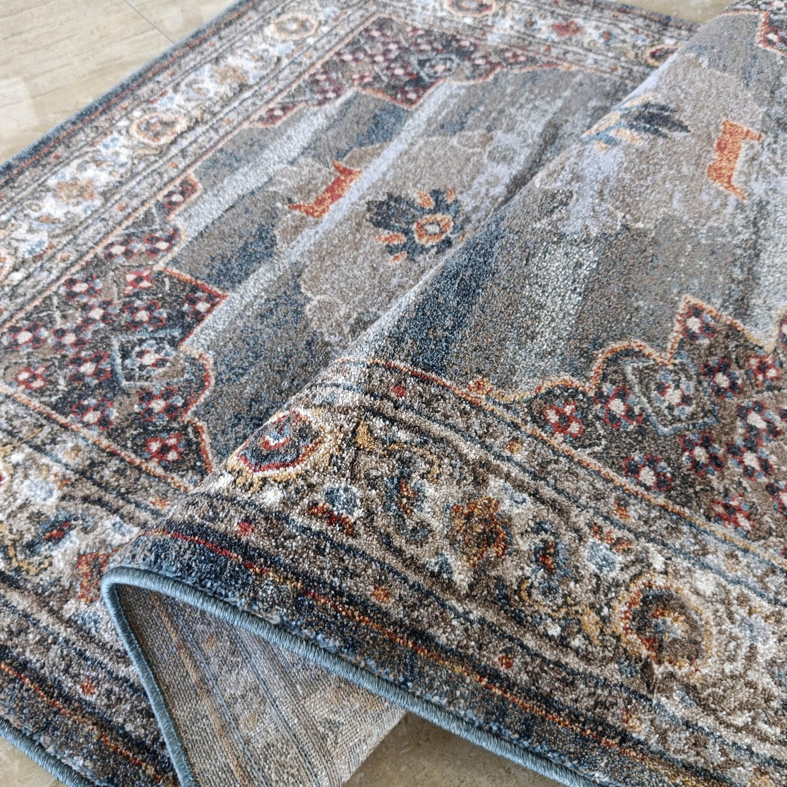 Tapis doux gris, bleu et ocre avec motifs traditionnels floraux et géométriques, partiellement enroulé sur un carrelage beige