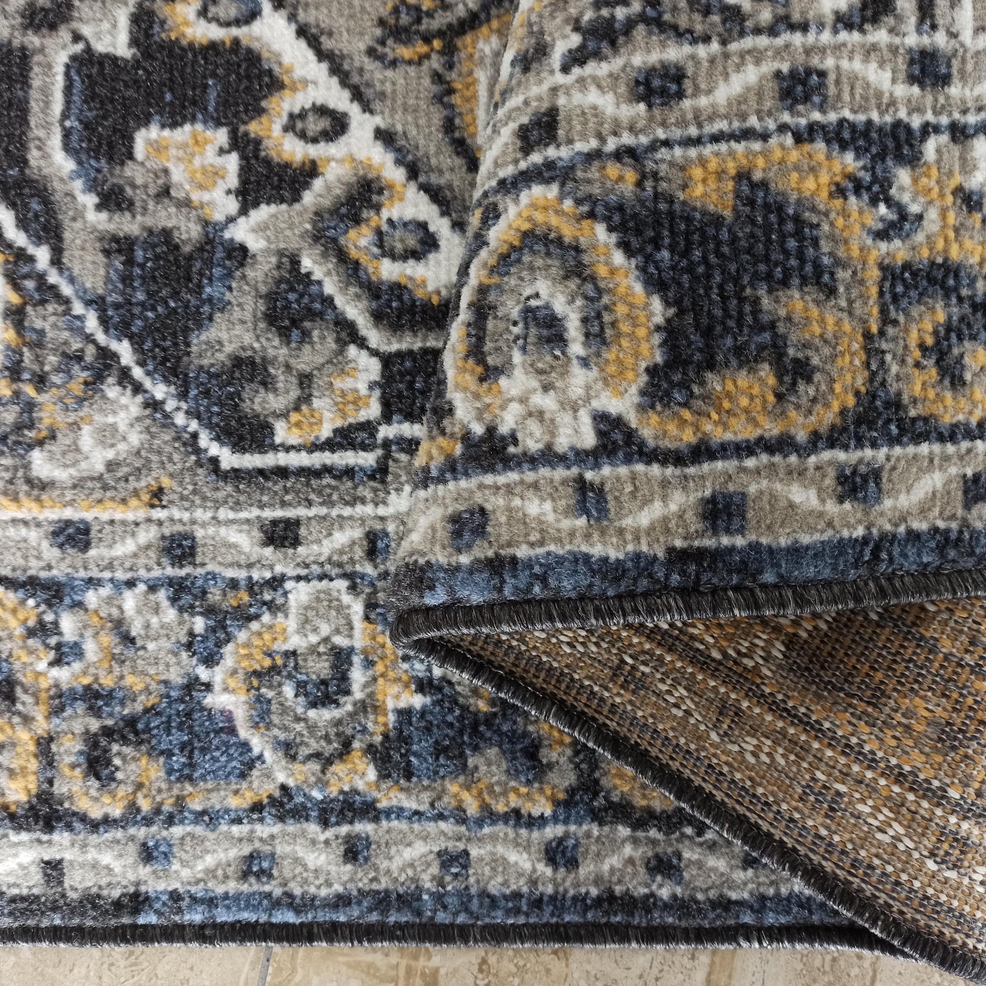 Tapis en textile doux aux motifs ornementaux gris, bleus, blancs et dorés, plié sur un sol carrelé