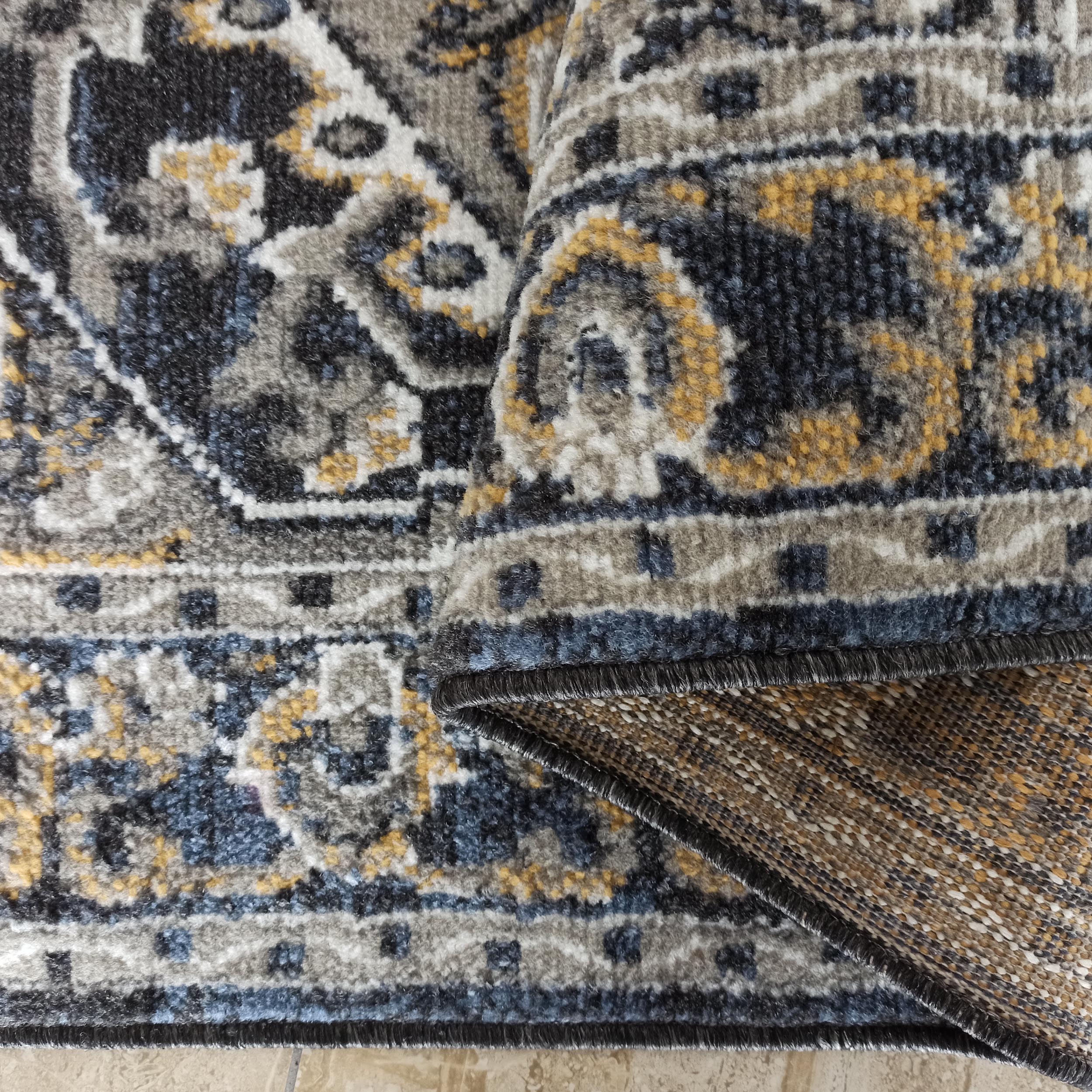 Tapis en textile doux aux motifs ornementaux gris, bleus, blancs et dorés, plié sur un sol carrelé