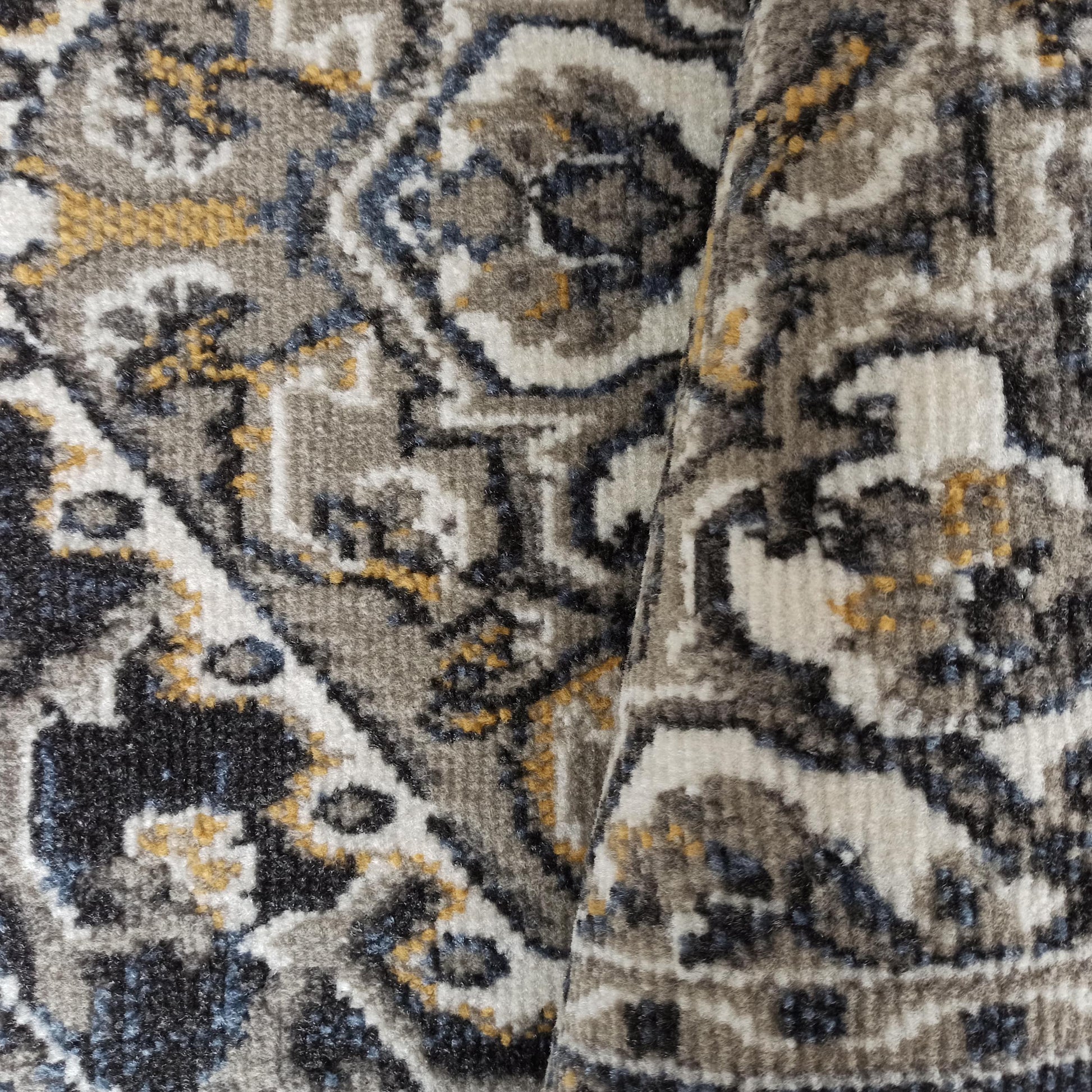 Tapis texturé à motifs abstraits complexes, aux couleurs gris, beige, blanc cassé, bleu ardoise et jaune doré, visiblement plié