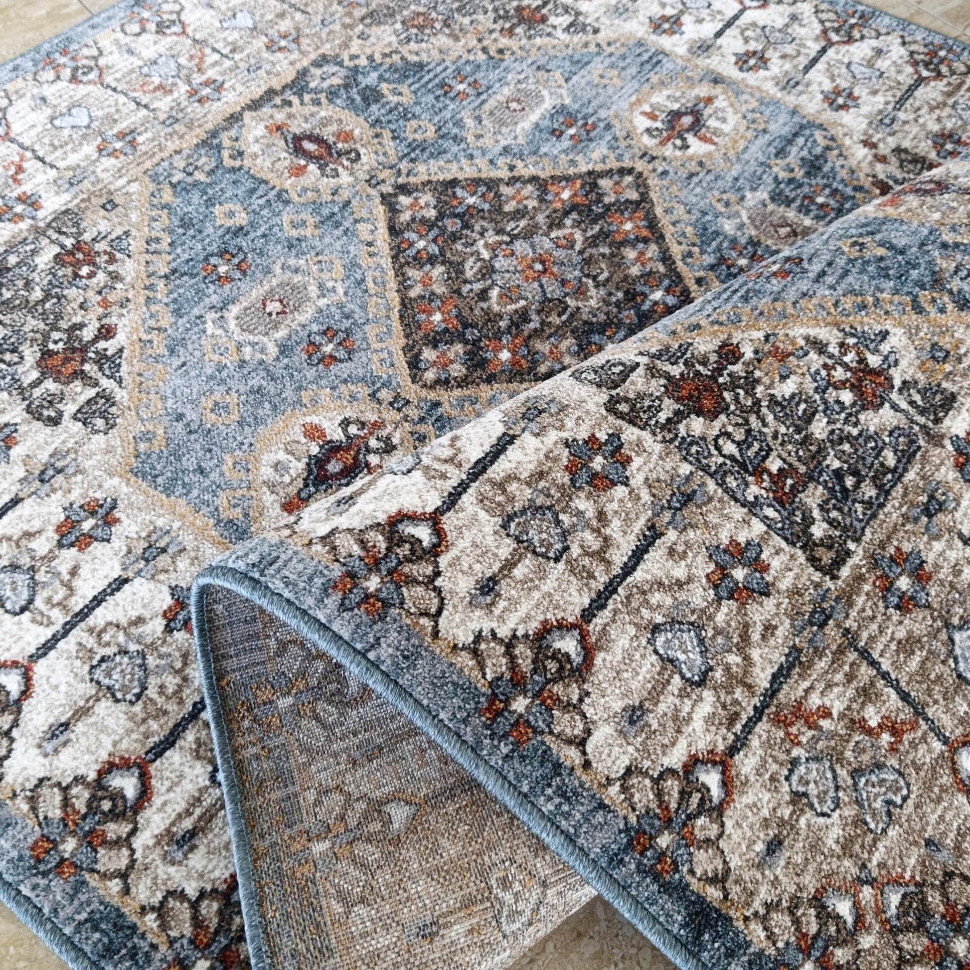 Tapis doux à motifs traditionnels bleu ardoise, crème, brun et orange sur sol carrelé clair, partiellement roulé