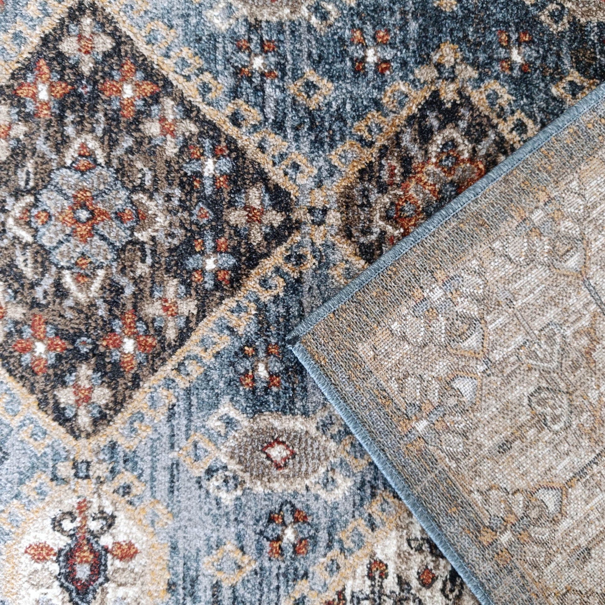 Tapis moelleux aux motifs bleus, marrons, noirs et orangés, avec dessous tissé beige visible et bord surjeté bleu