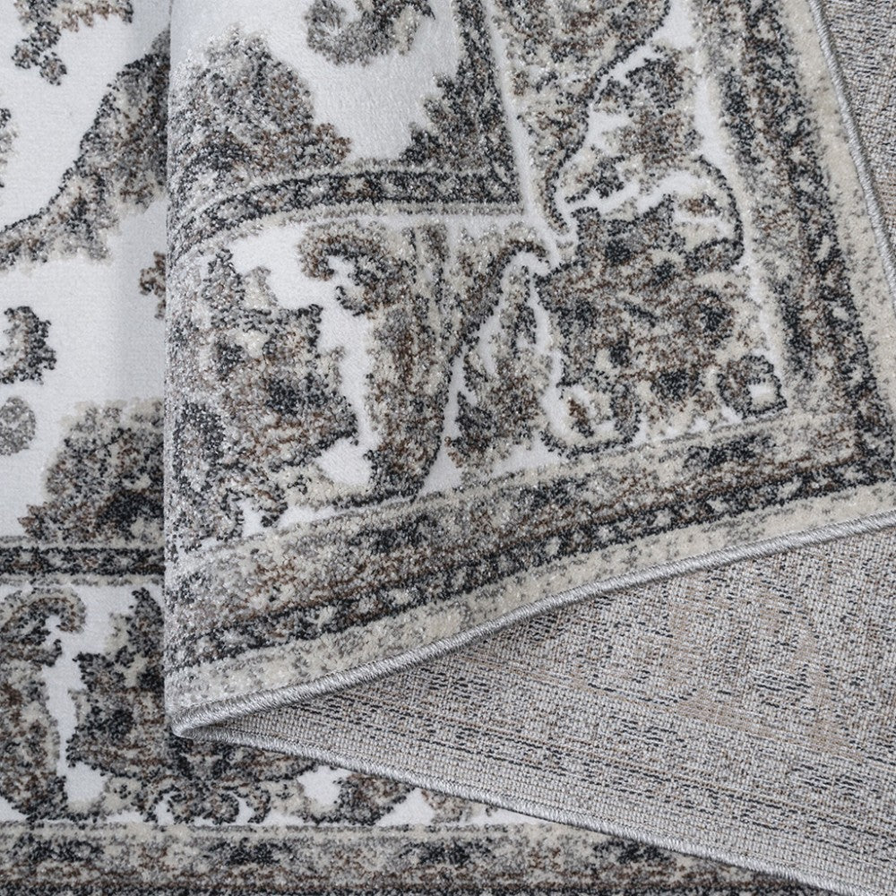 Tapis moelleux aux motifs ornementaux beige et gris-noir sur fond blanc, une bordure visible et un pli expose son envers tissé