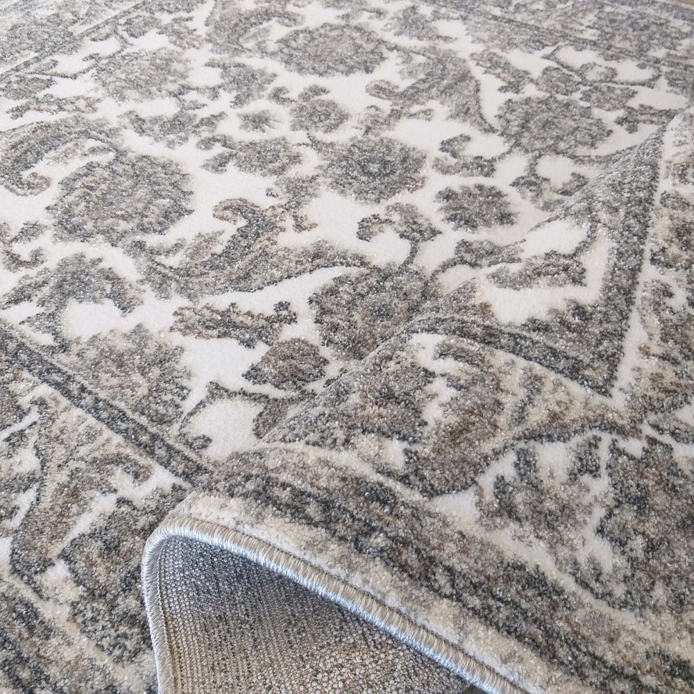 Tapis texturé crème et gris aux motifs ornementaux, avec un coin relevé dévoilant son envers textile