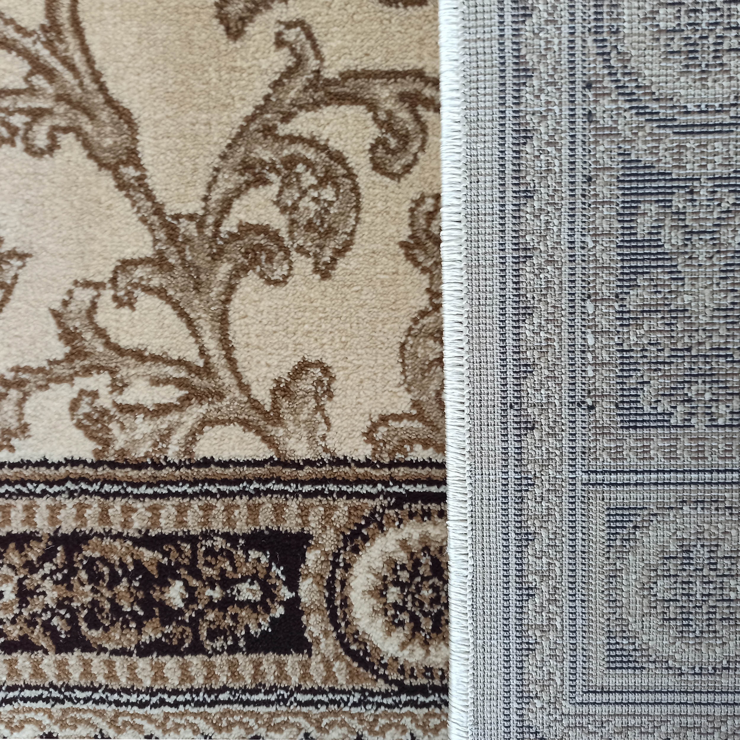 Tapis beige et marron à motifs floraux et bordure ornementale, partiellement roulé pour montrer son envers tissé grisâtre