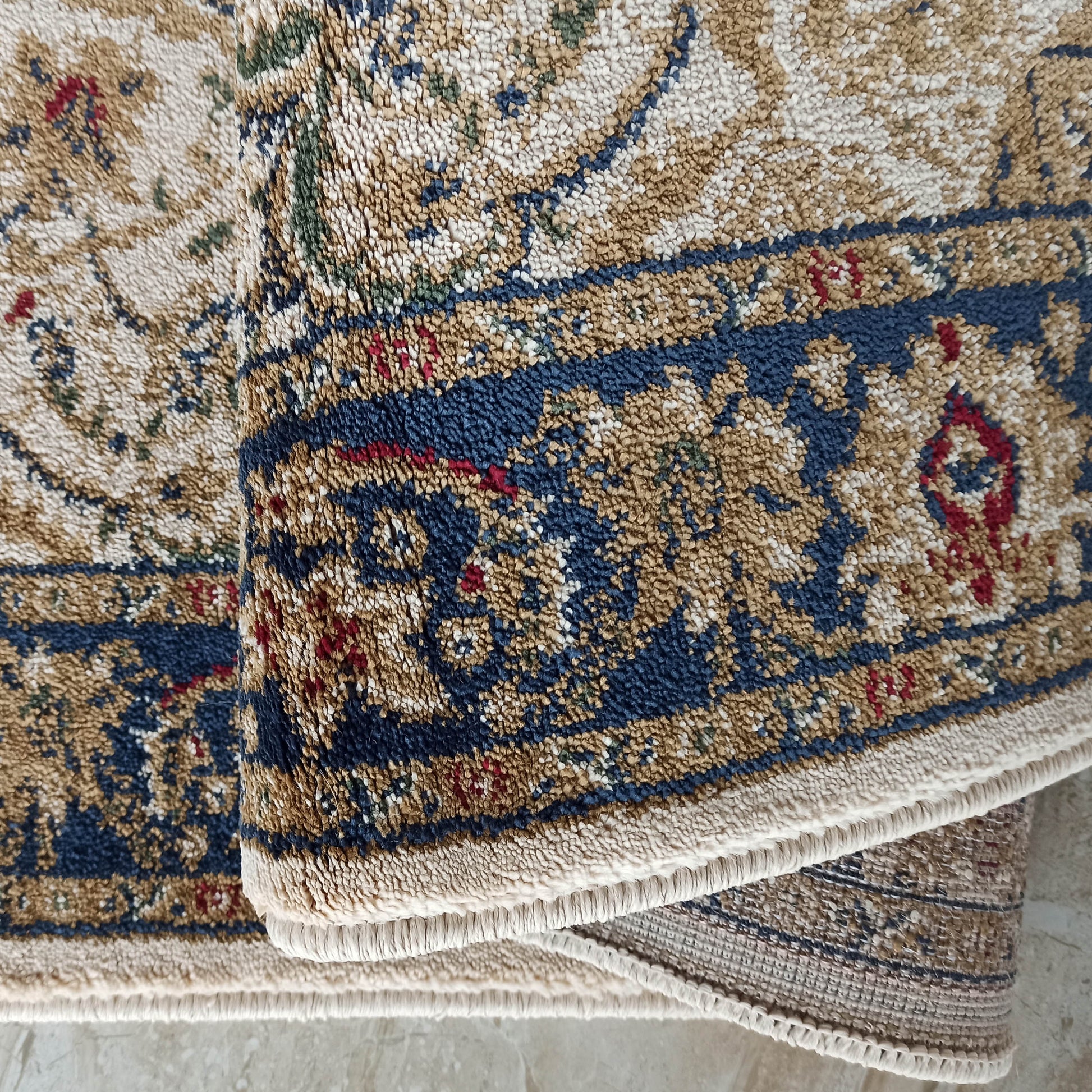 Gros plan sur un tapis texturé aux motifs traditionnels beiges, bleus foncés, rouges et verts, partiellement roulé