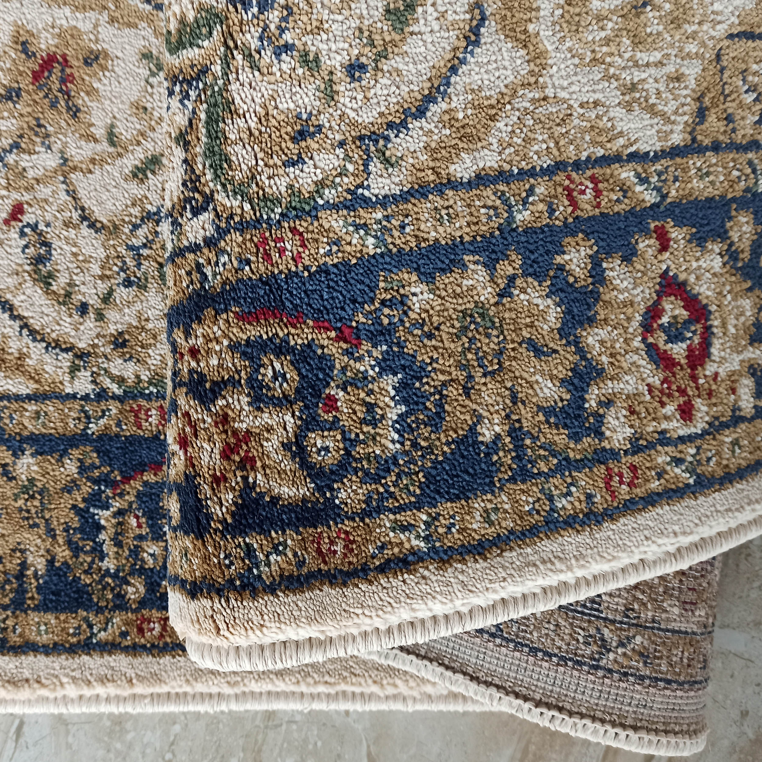 Gros plan sur un tapis texturé aux motifs traditionnels beiges, bleus foncés, rouges et verts, partiellement roulé