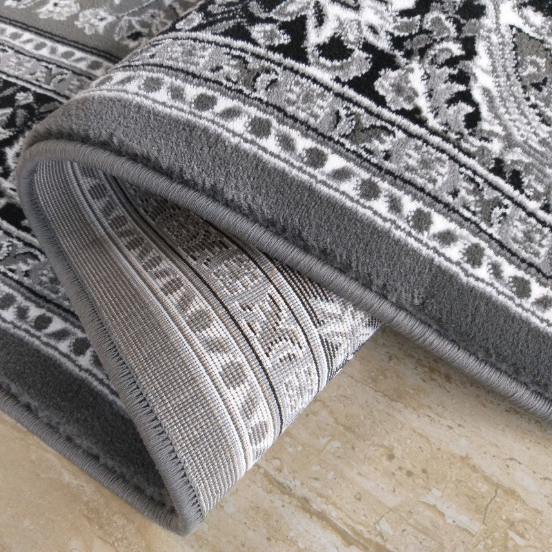 Tapis gris, noir et blanc avec motifs ornementaux, partiellement roulé pour montrer son envers tissé géométrique