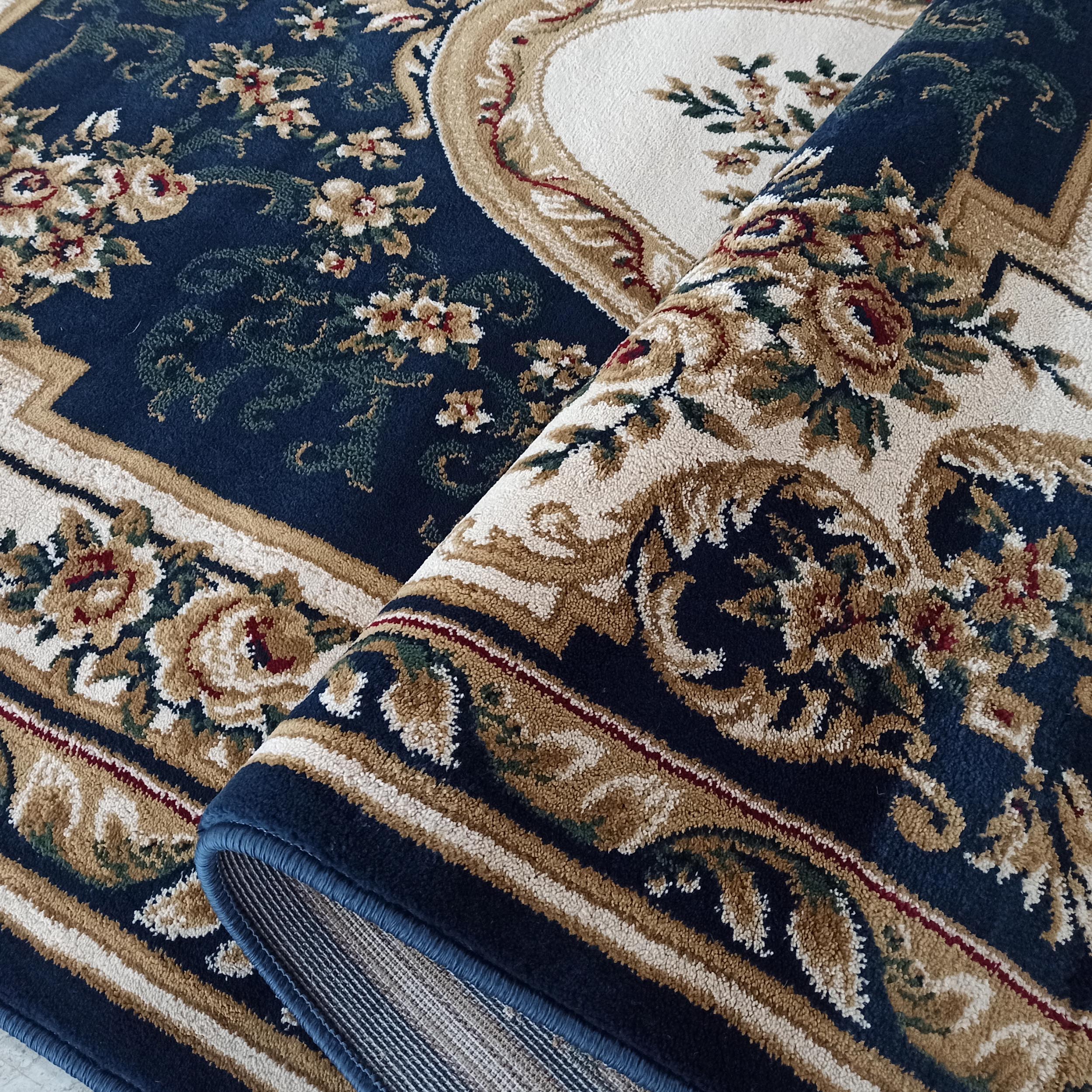 Tapis à motifs floraux complexes en bleu marine, beige, doré et rouge, partiellement roulé révélant sa texture douce