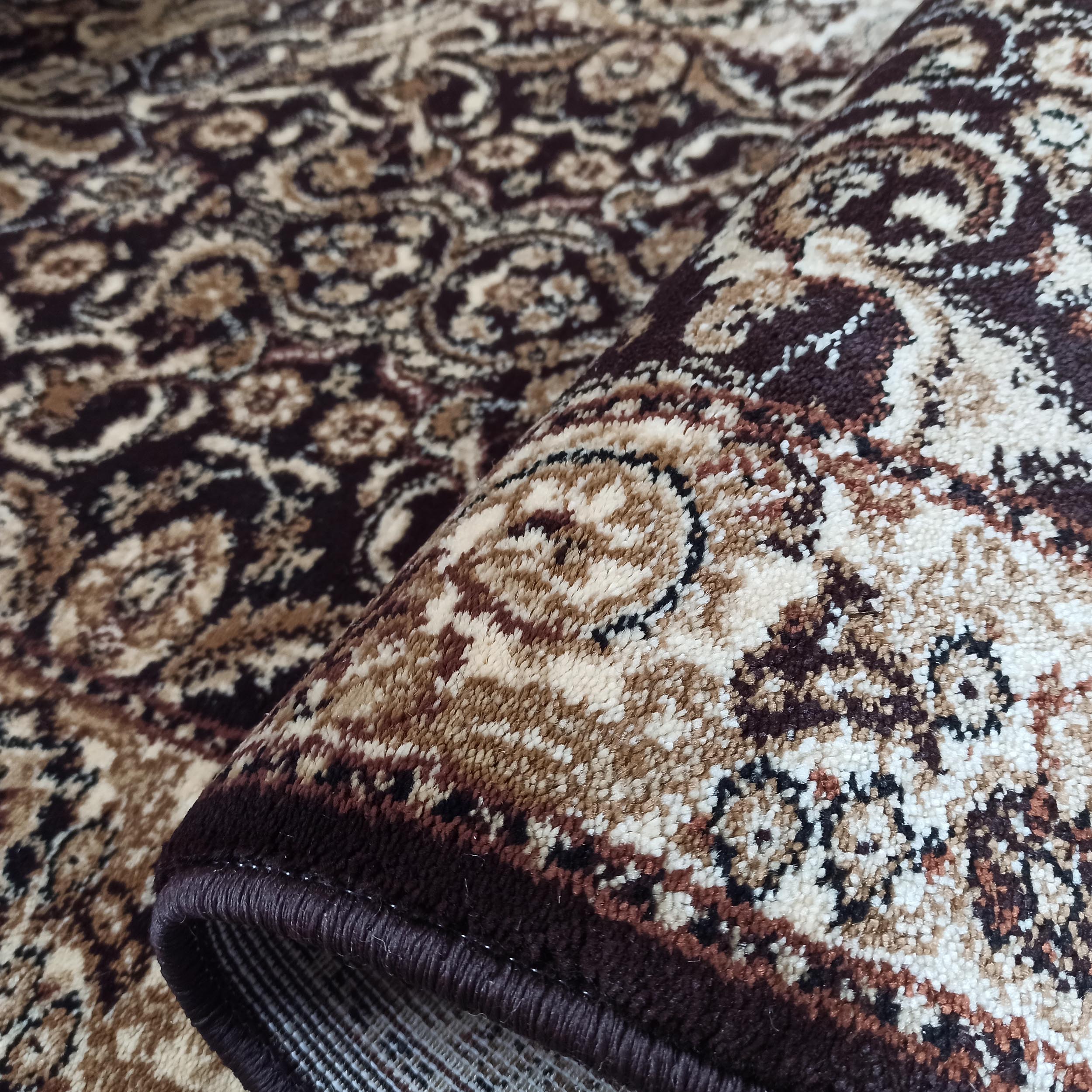Tapis enroulé avec des motifs ornementaux complexes marron foncé, beige et crème. La texture du velours est visible