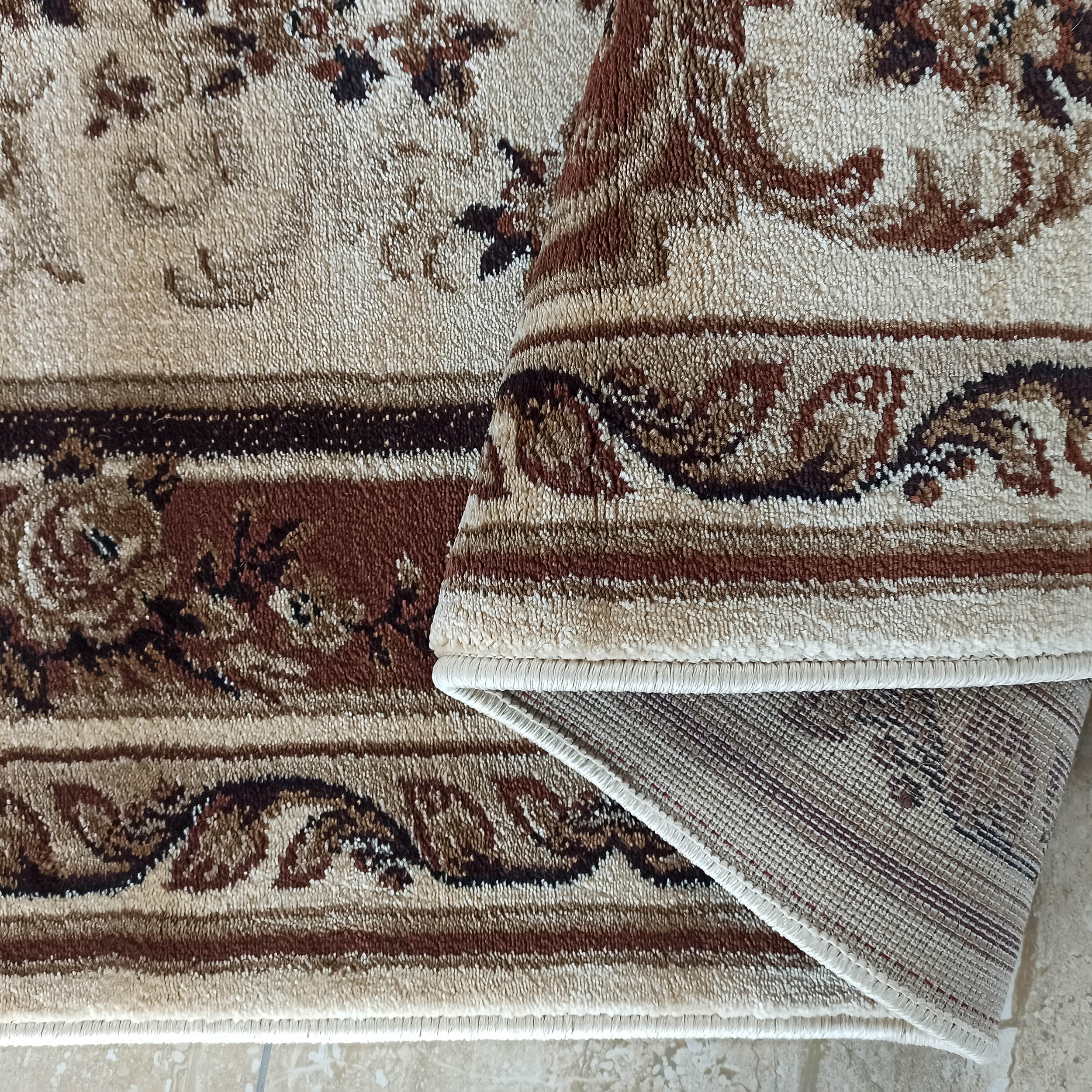 Tapis beige et marron aux motifs floraux ornementaux, dont une partie est roulée, montrant le dessous du textile