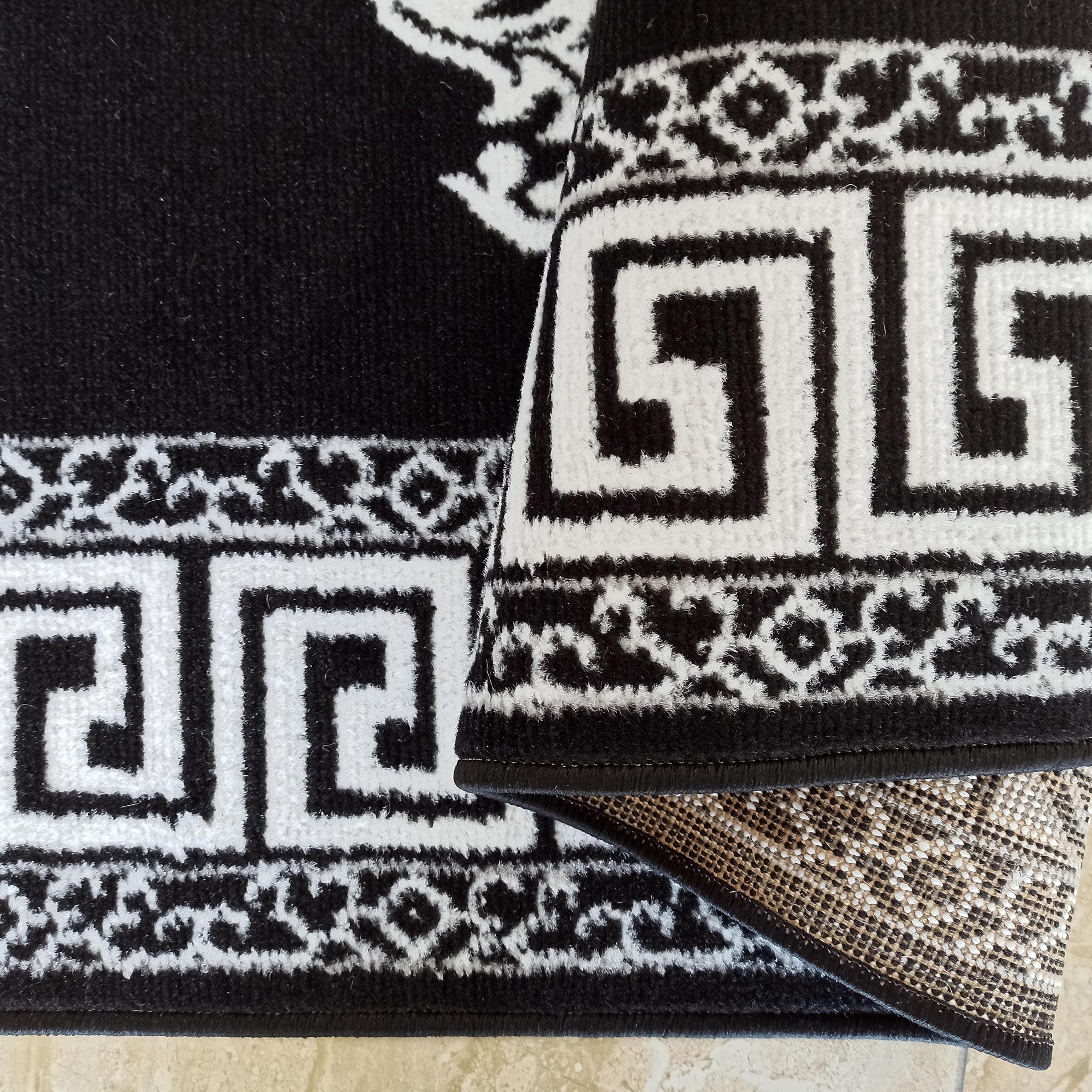 Tapis noir et blanc doux avec motifs de méandre et entrelacés, une section enroulée montre son envers texturé beige