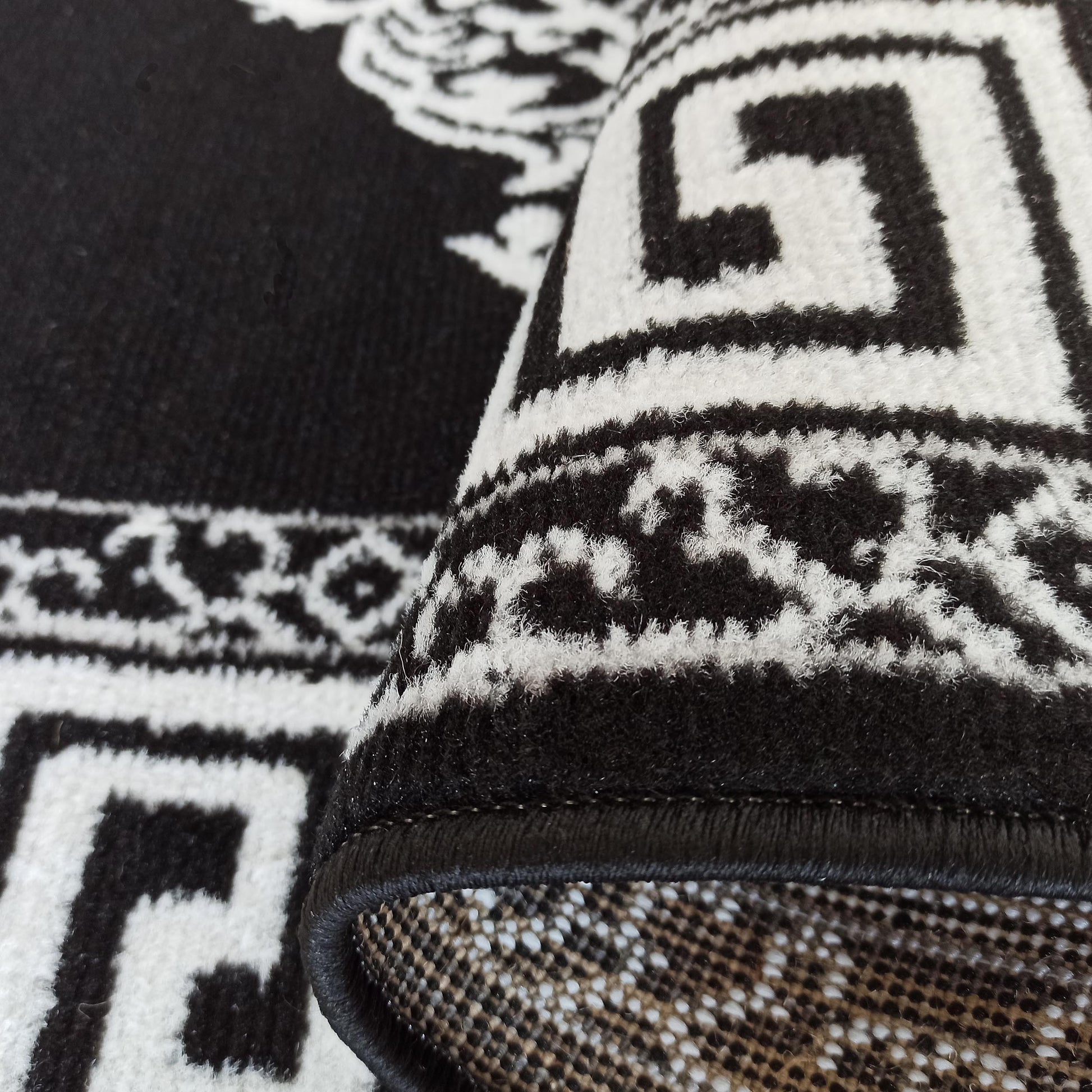 Tapis noir et blanc à motifs de méandre et ornements stylisés, texture douce, son bord roulé révèle le tissage du dessous