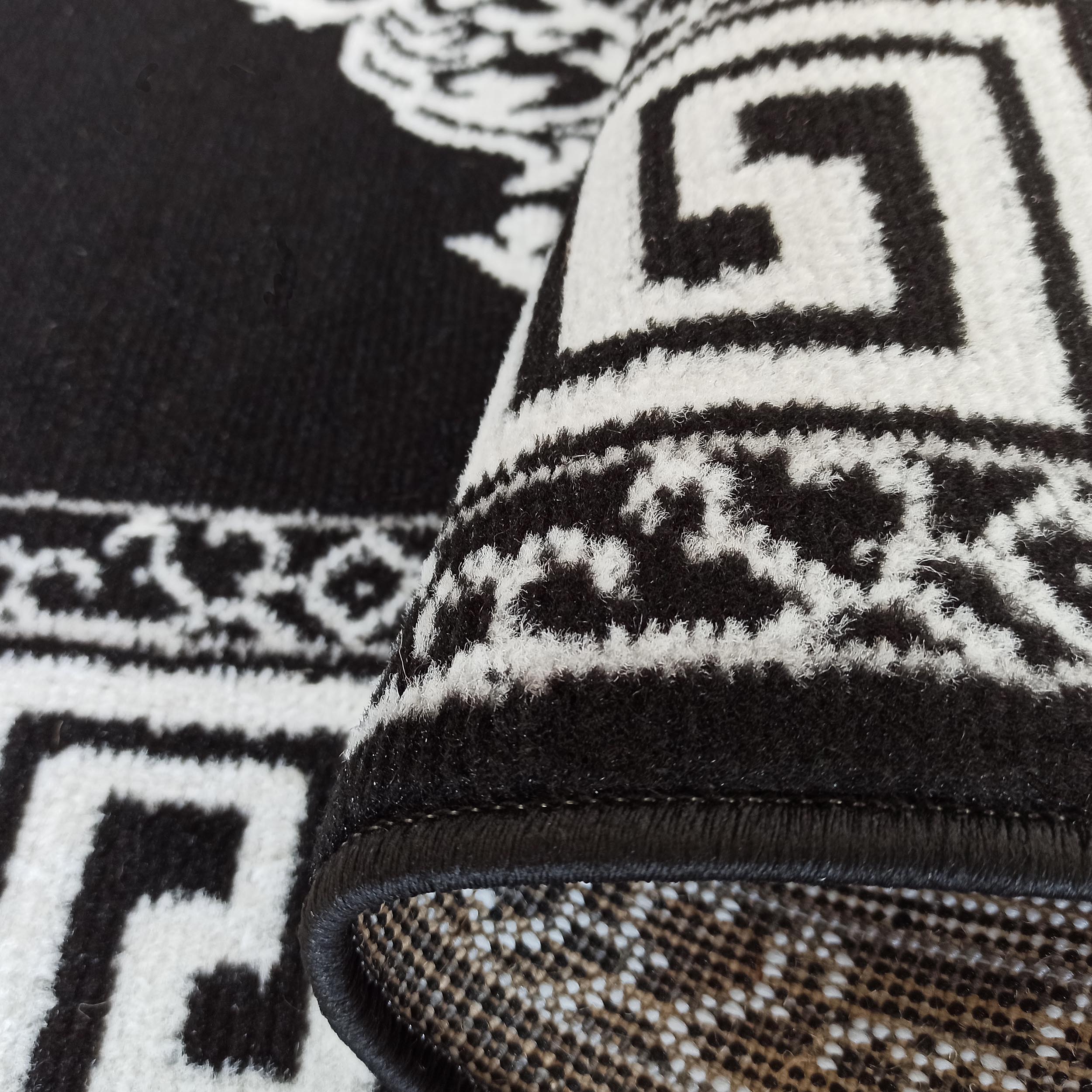 Tapis noir et blanc à motifs de méandre et ornements stylisés, texture douce, son bord roulé révèle le tissage du dessous
