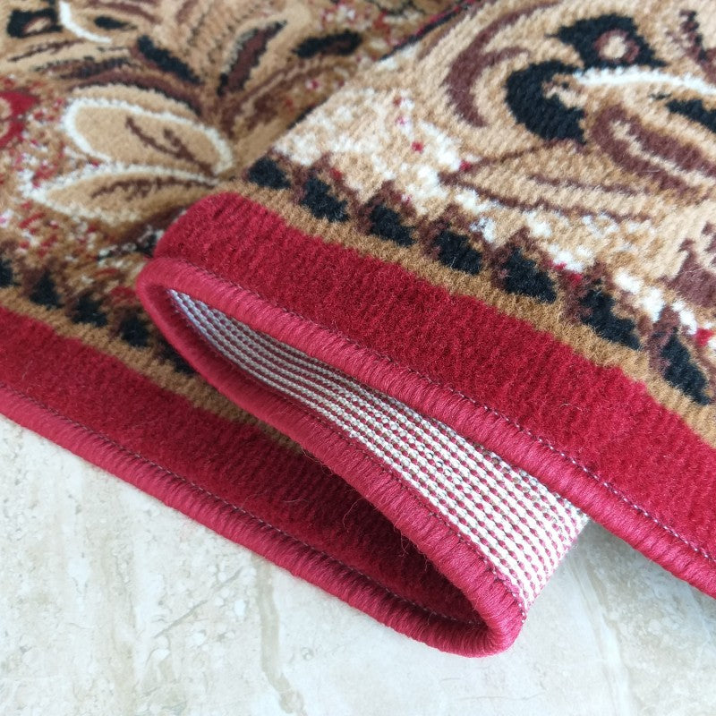 Tapis avec motifs ornementaux beige, marron et noir rehaussés de rouge. Le tapis est plié, montrant sa bordure rouge et son envers tissé