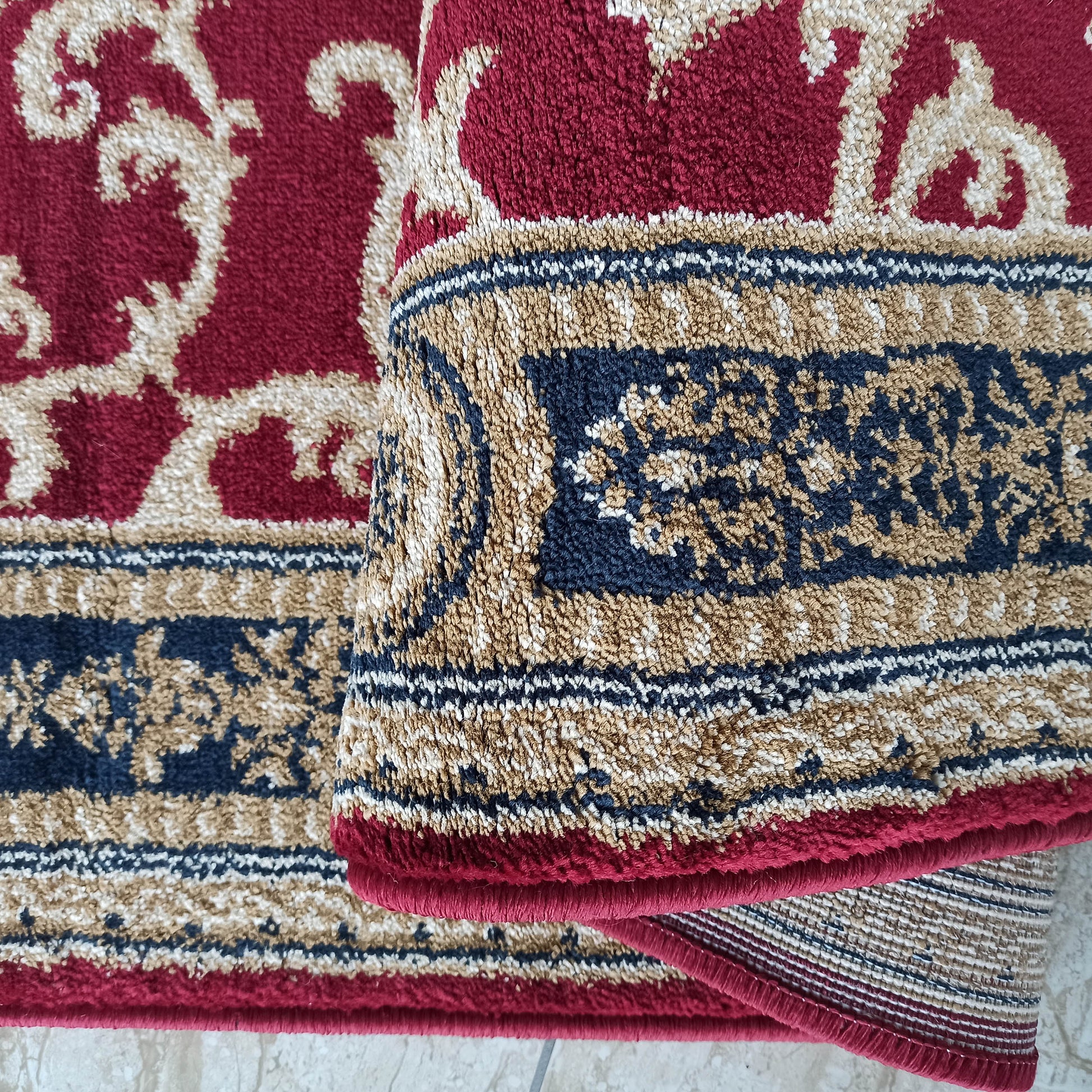 Tapis rouge bordeaux aux motifs arabesques beiges et à la bordure bleue et dorée Sa texture épaisse révèle un envers tissé