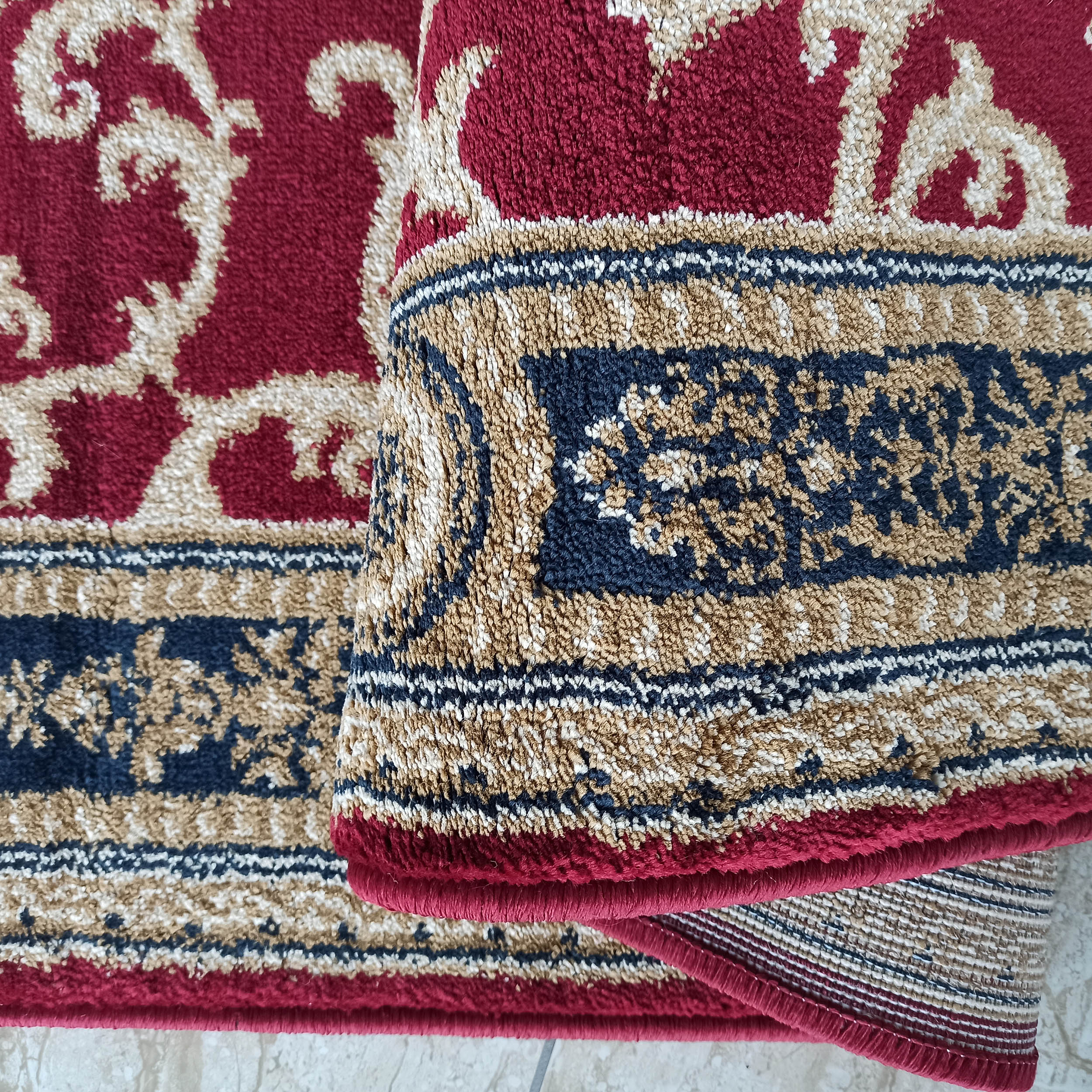 Tapis rouge bordeaux aux motifs arabesques beiges et à la bordure bleue et dorée Sa texture épaisse révèle un envers tissé