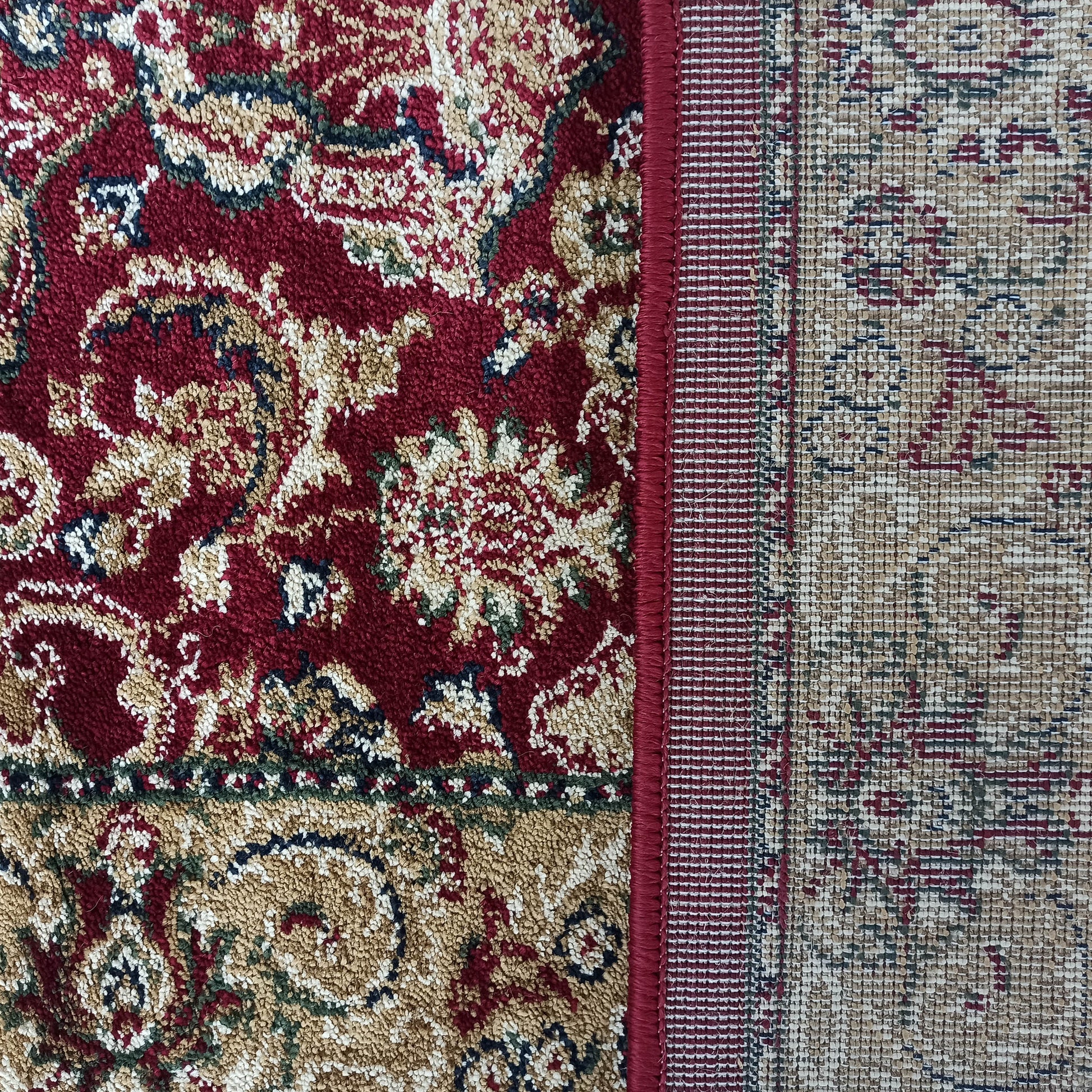 Tapis bordeaux orné de motifs floraux dorés, bleus et verts. Le revers tissé et le surbord rouge sont visibles