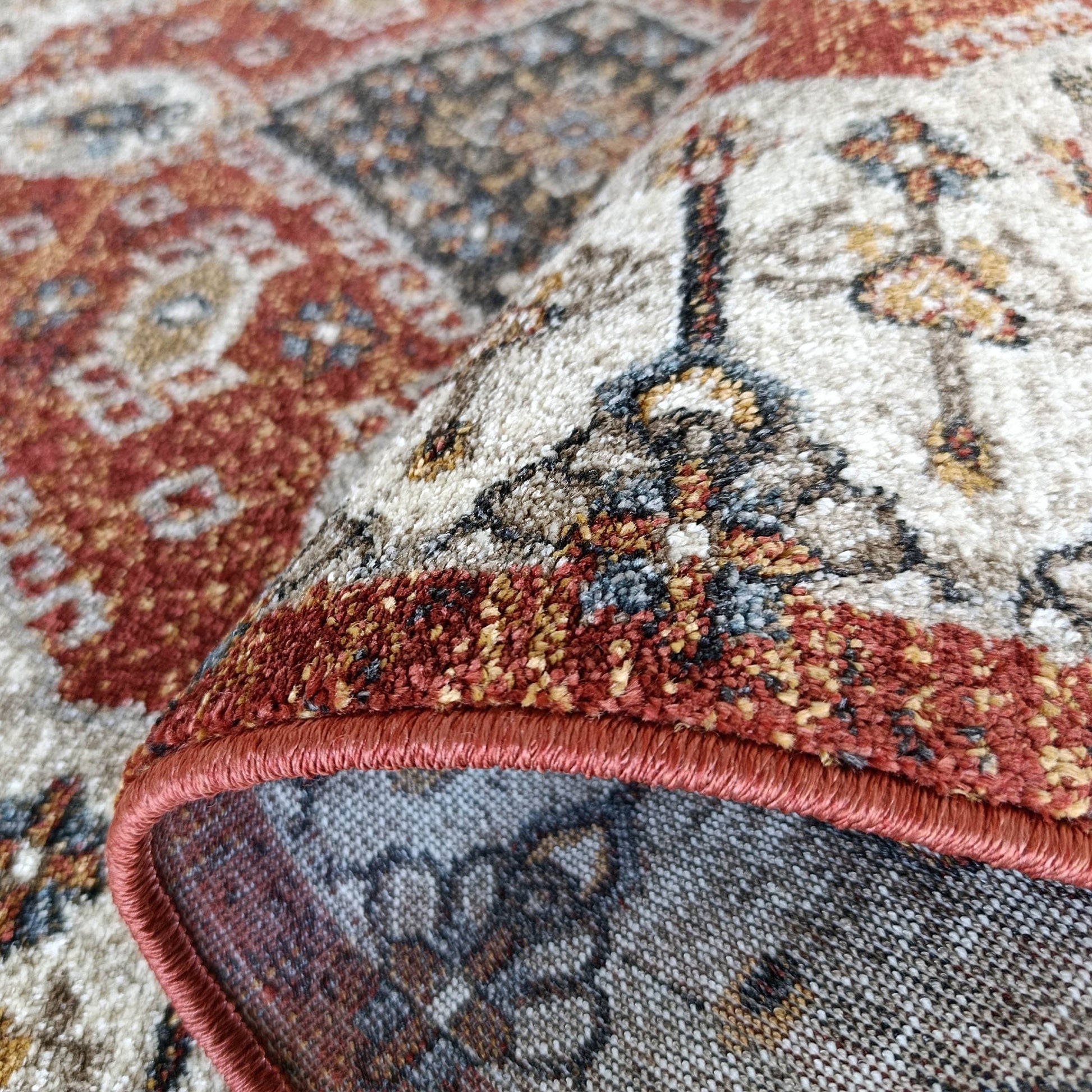 Un tapis enroulé avec des motifs complexes en rouge terre cuite, crème, gris et bleu, sa surface douce et son bord surjeté