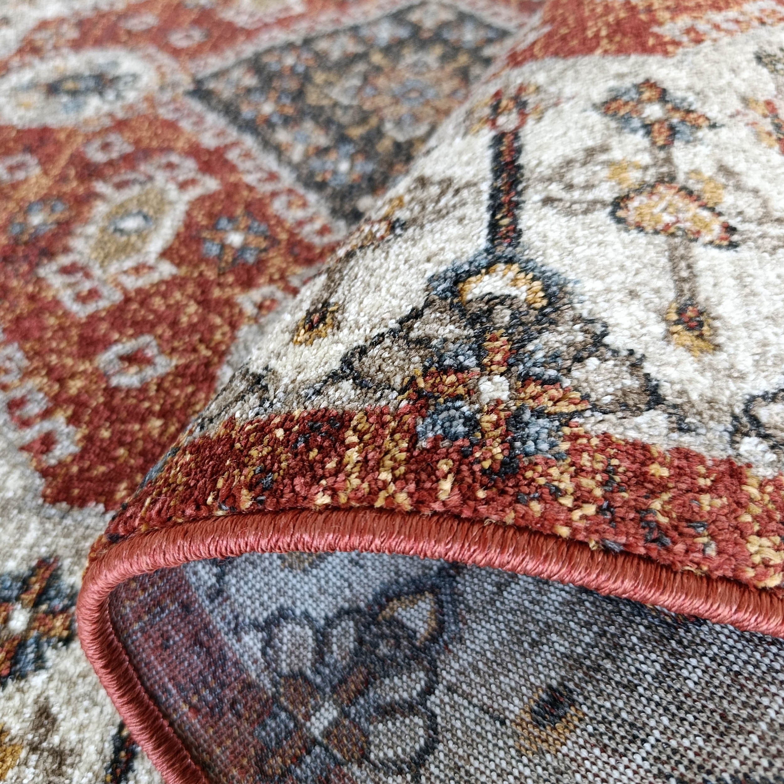 Un tapis enroulé avec des motifs complexes en rouge terre cuite, crème, gris et bleu, sa surface douce et son bord surjeté