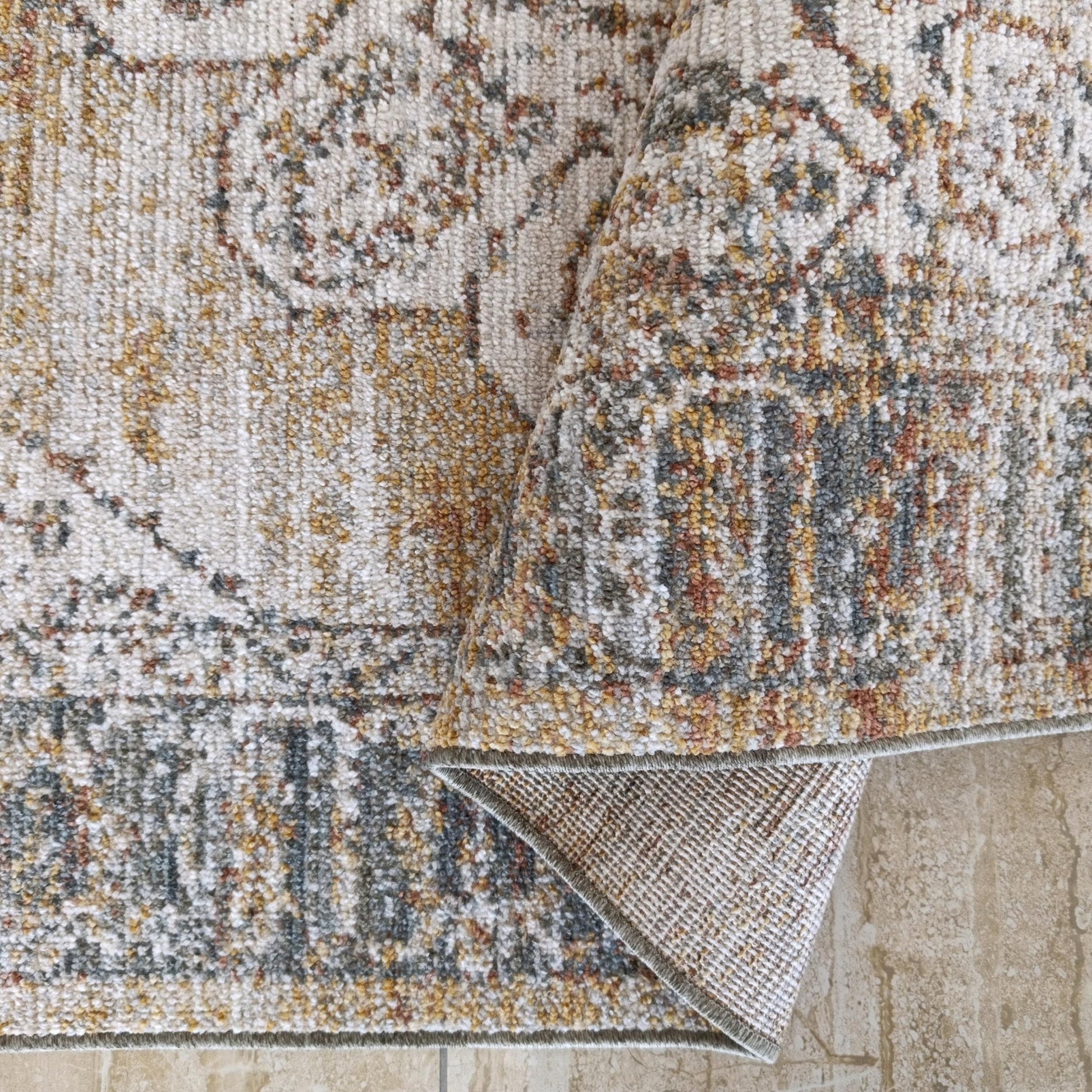 Tapis à motifs entrelacés crème, gris, ocre et rouille, son envers tissé apparent sur un sol carrelé beige
