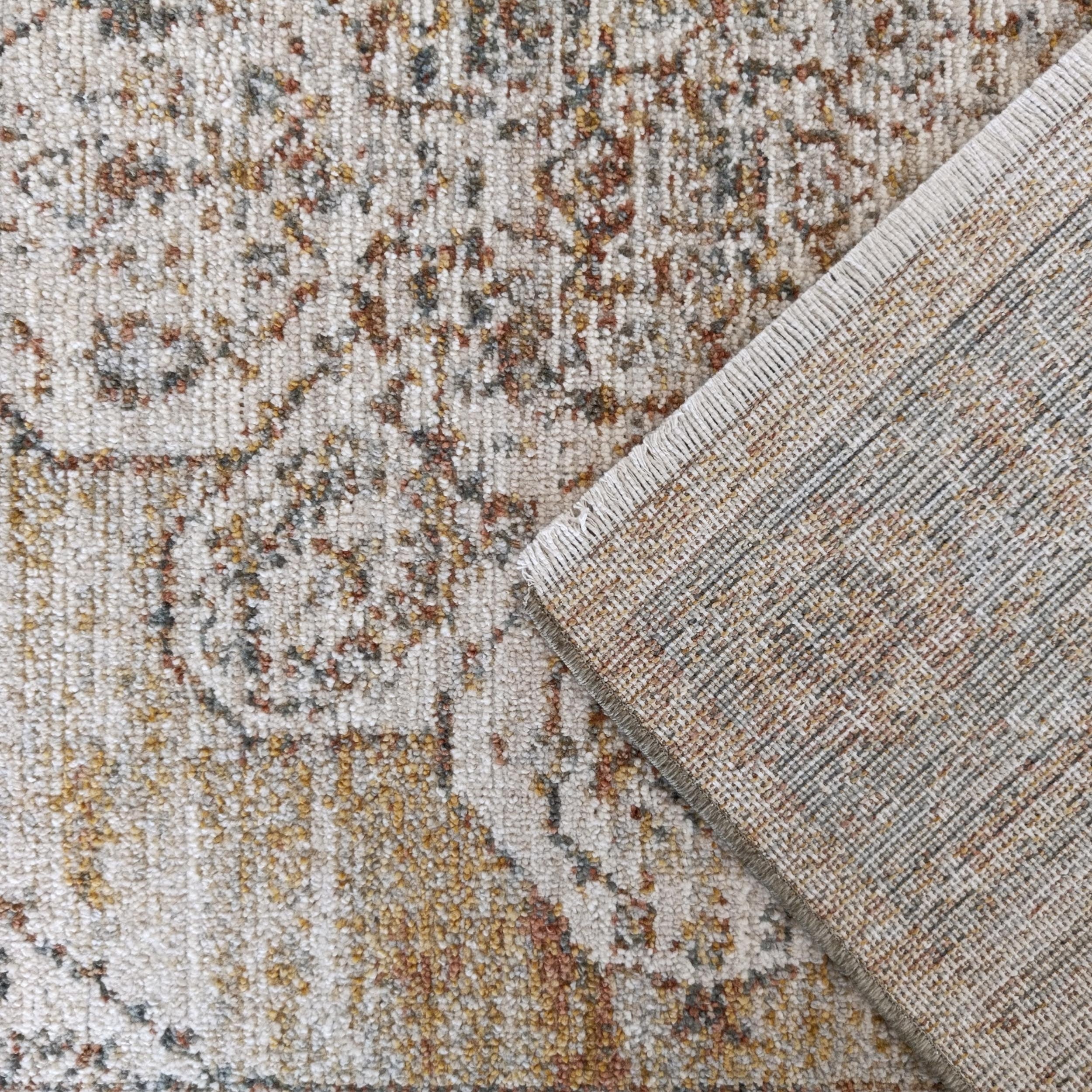Tapis aux motifs clairs et vieillis beige, rouille et ocre, partiellement replié, dévoilant son envers en tissu tissé gris