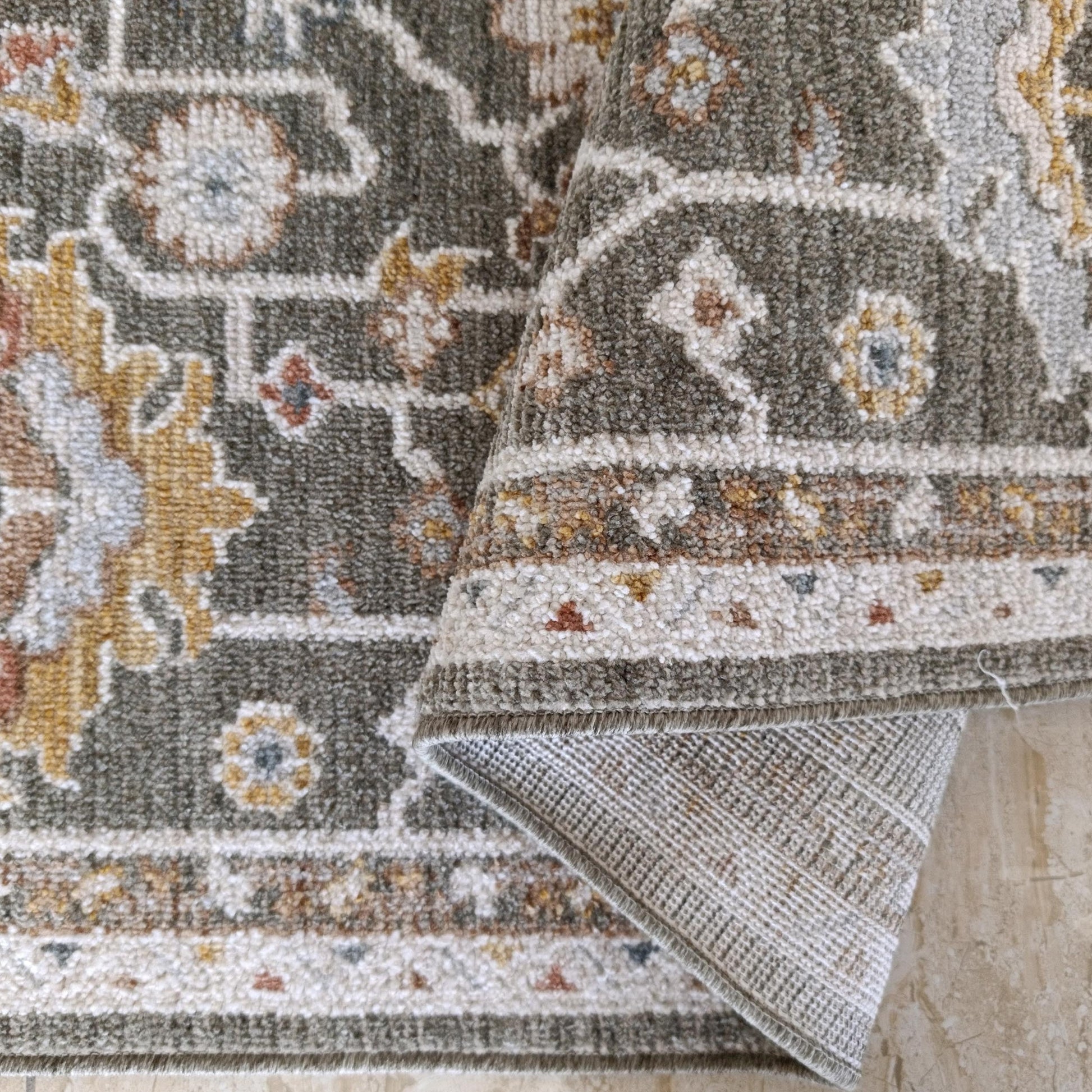 Tapis tissé à motifs floraux et géométriques kaki, crème, doré, rouille et bleu clair, partiellement enroulé sur un sol clair