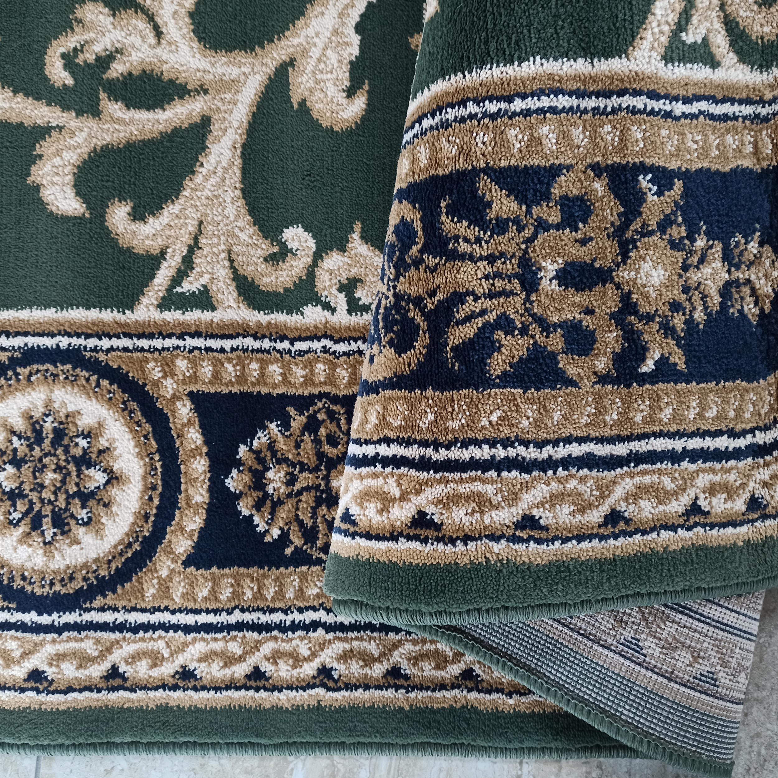 Tapis vert foncé aux motifs de volutes beiges, avec des bordures à motifs floraux et géométriques bleus, beiges et blancs, une partie est pliée