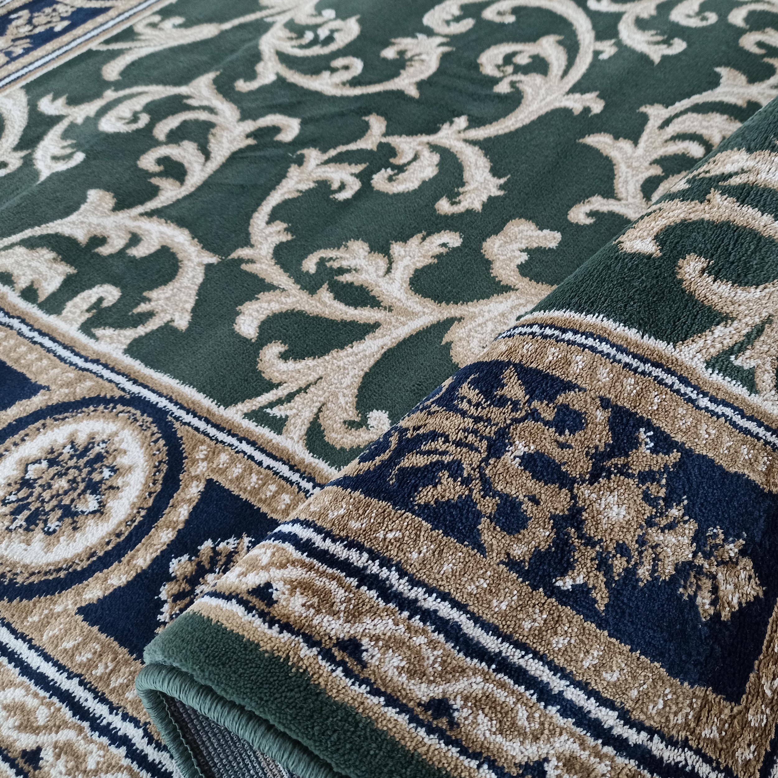 Tapis vert forêt avec motifs arabesques crème, bordure en bandes bleu marine, beige et blanc, visiblement moelleux et enroulé