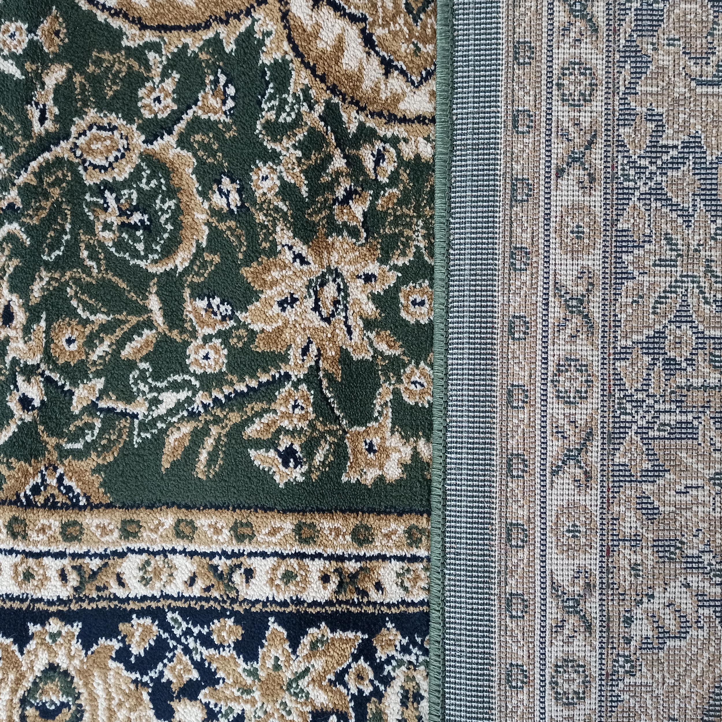 Tapis avec motifs ornementaux beiges, noirs et crème sur fond vert foncé, l'envers tissé beige et gris apparaît à droite