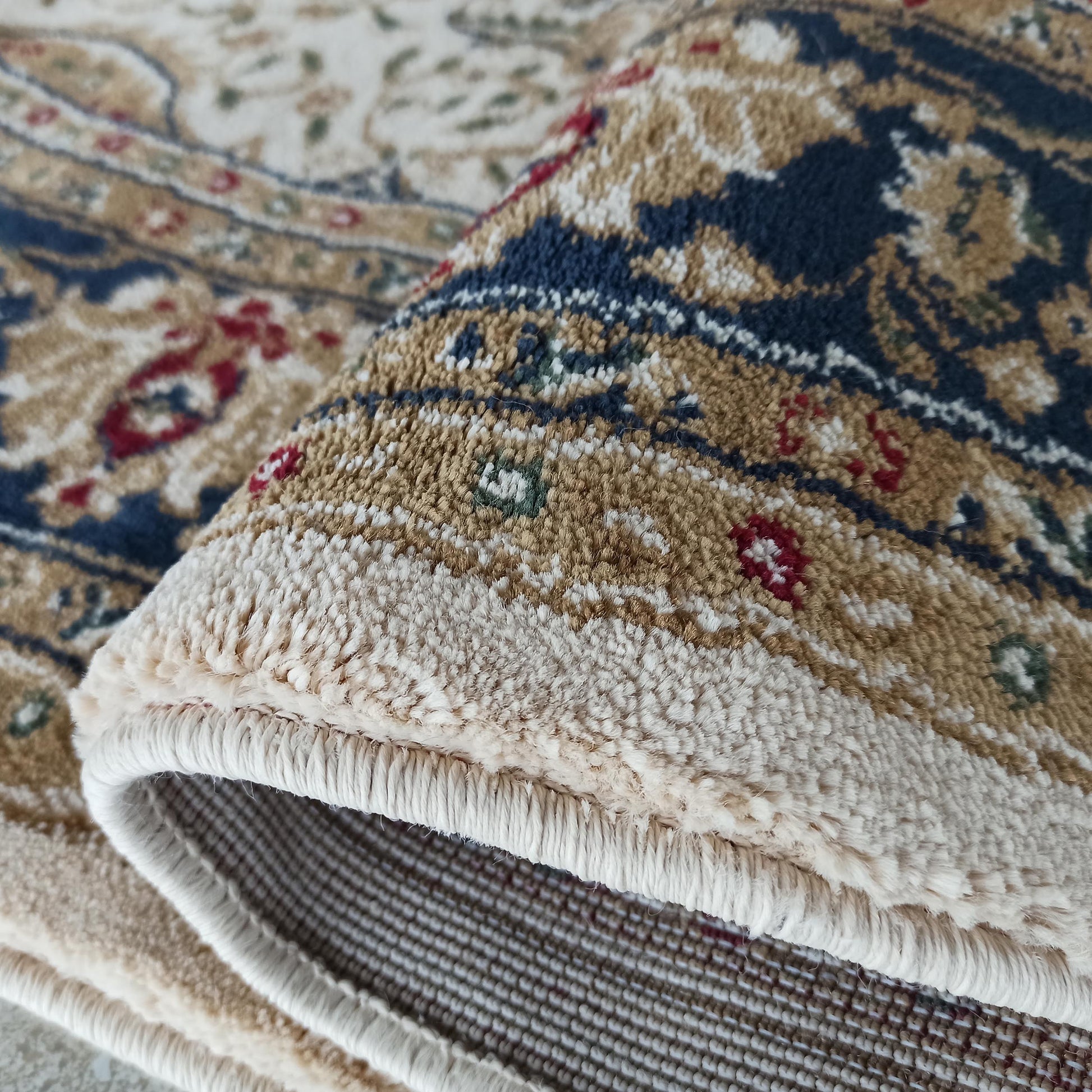 Tapis ornemental aux teintes beiges, bleues, dorées et rouges, montrant sa surface touffue et son revers tissé