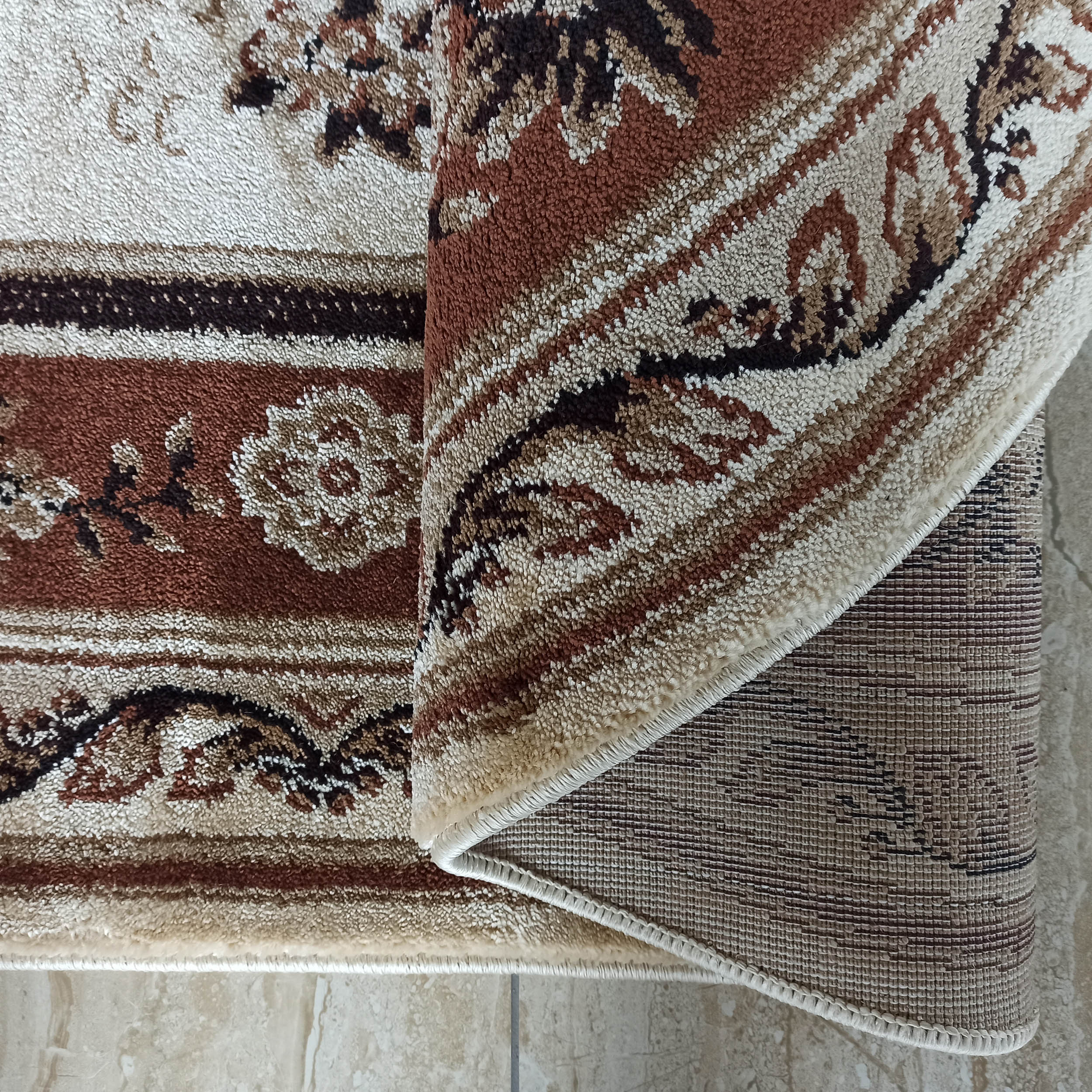 Tapis texturé beige, marron et crème aux motifs floraux complexes, avec l'envers tissé apparent car partiellement enroulé