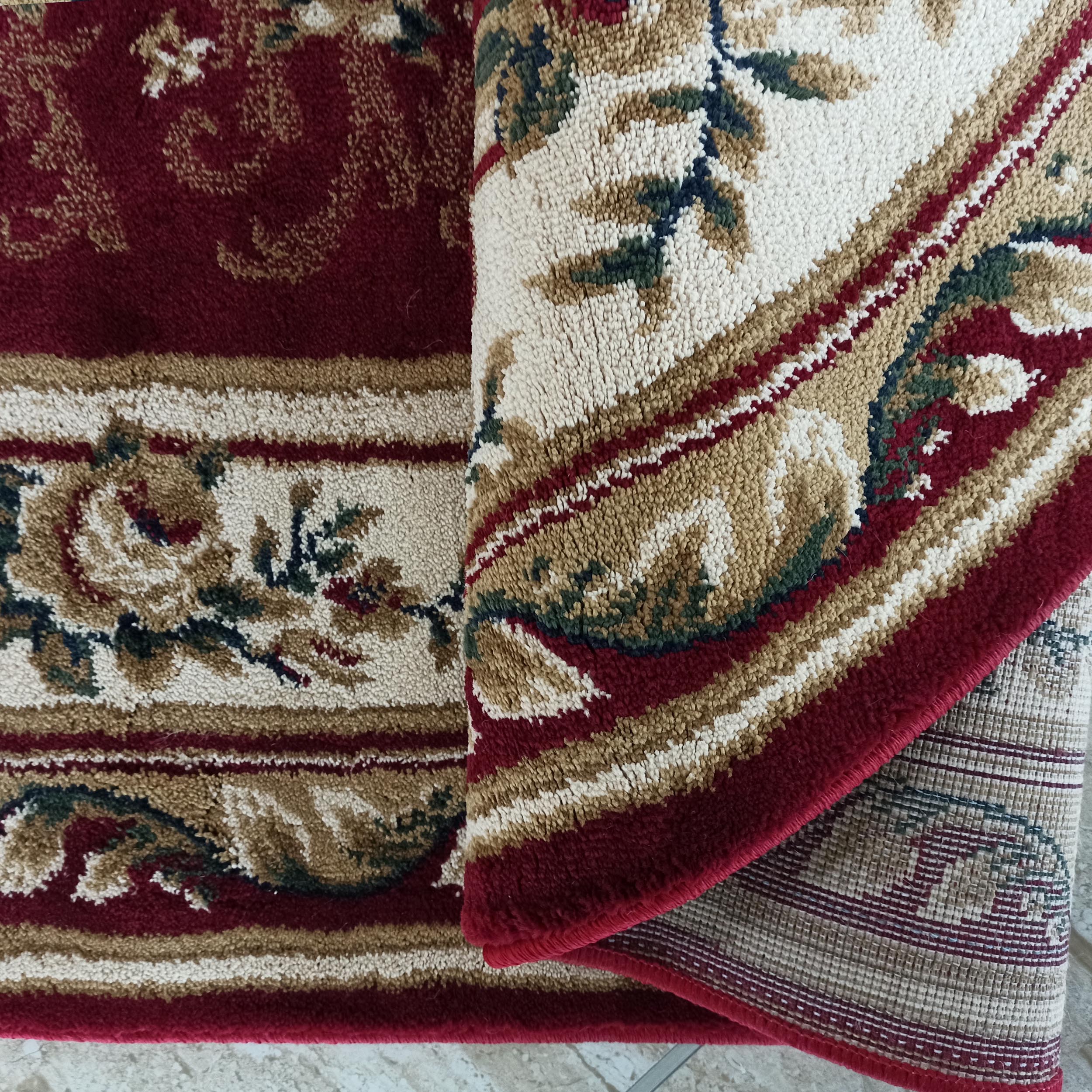 Tapis bordeaux à motifs floraux crème, beiges, verts et bleus. Une partie roulée révèle l'envers tissé à rayures