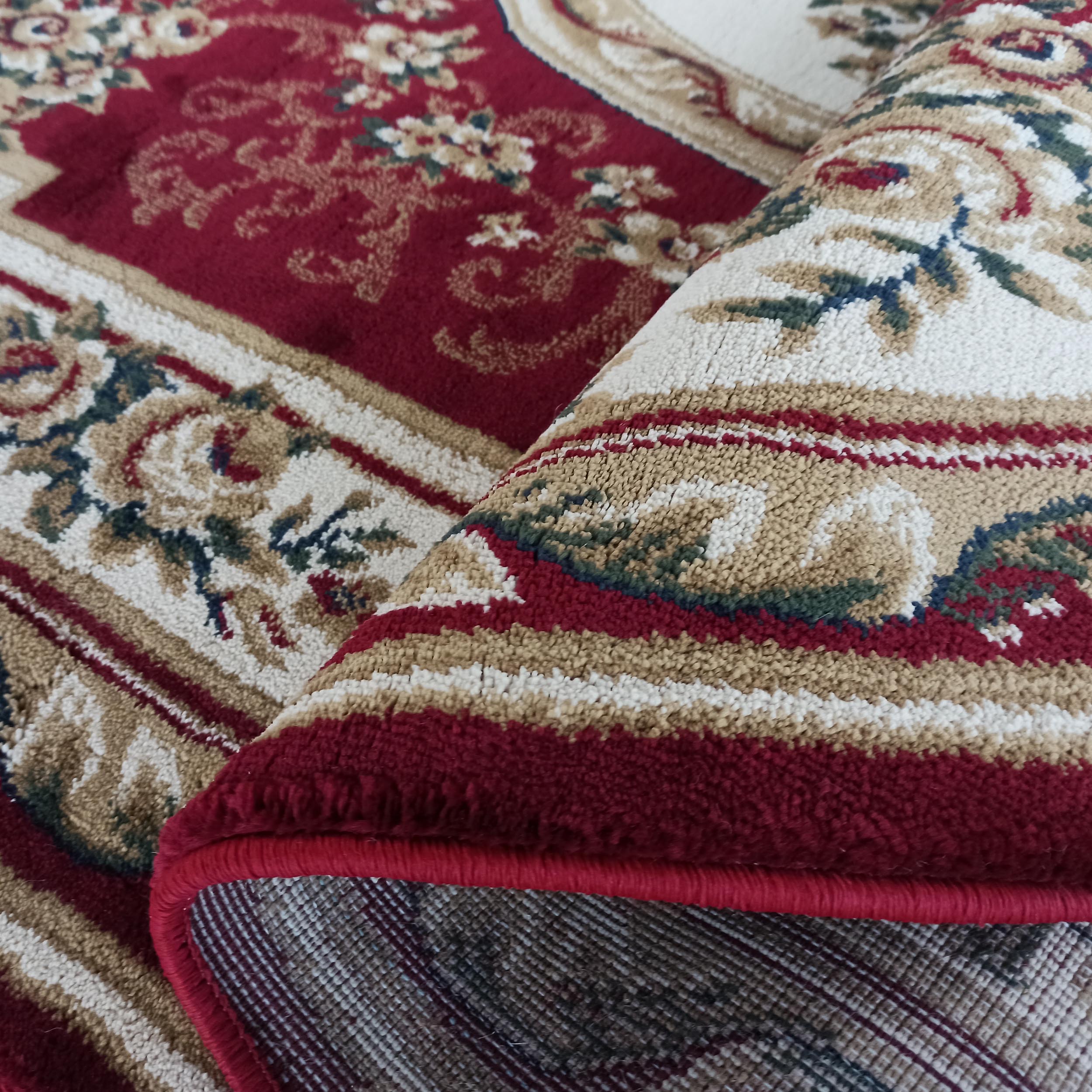 Un tapis rouge bordeaux à motifs floraux et arabesques crèmes, beiges et verts, dont une section est roulée montrant l'envers tissé
