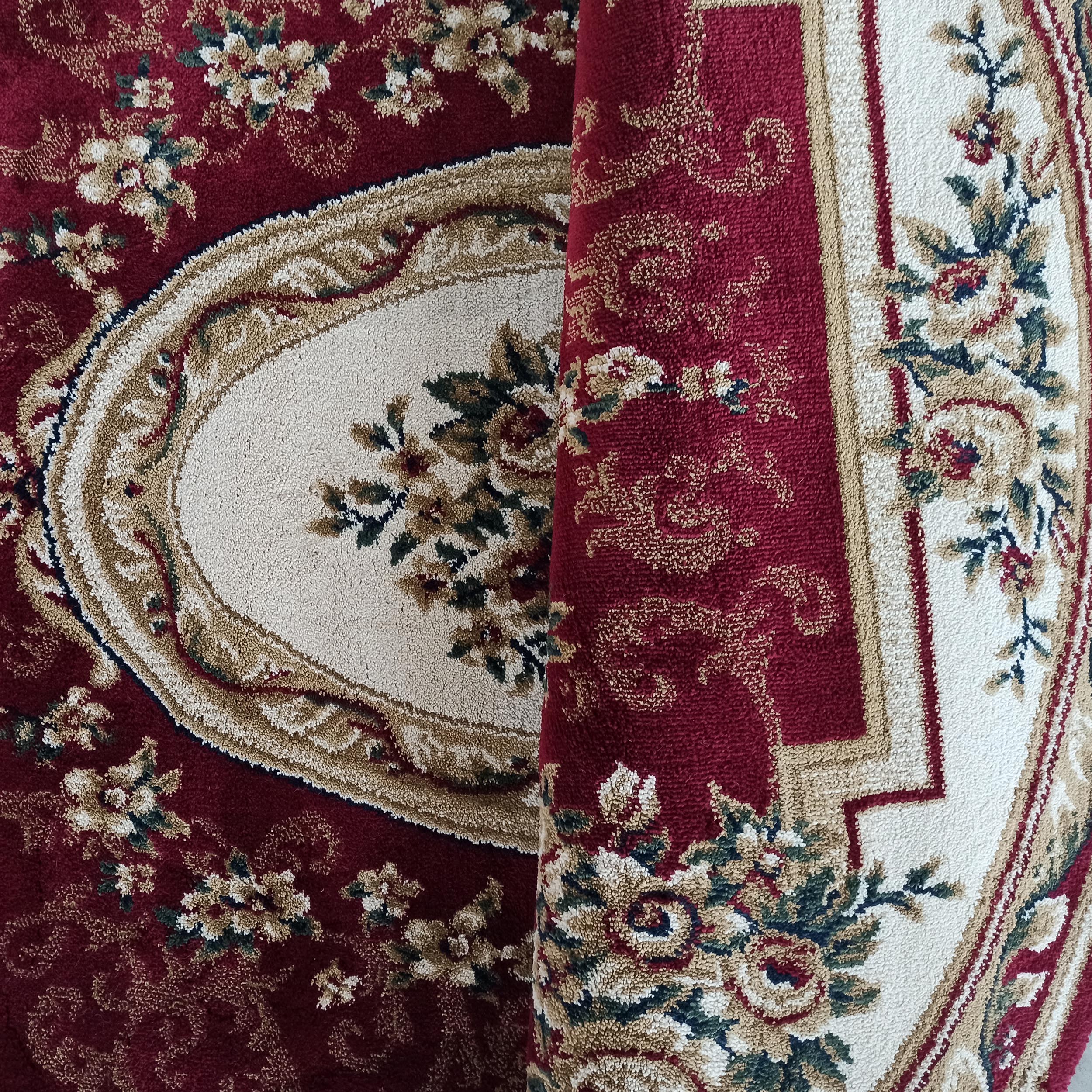 Tapis bordeaux orné de motifs floraux crème, or et verts, avec un médaillon central ovale, partiellement roulé
