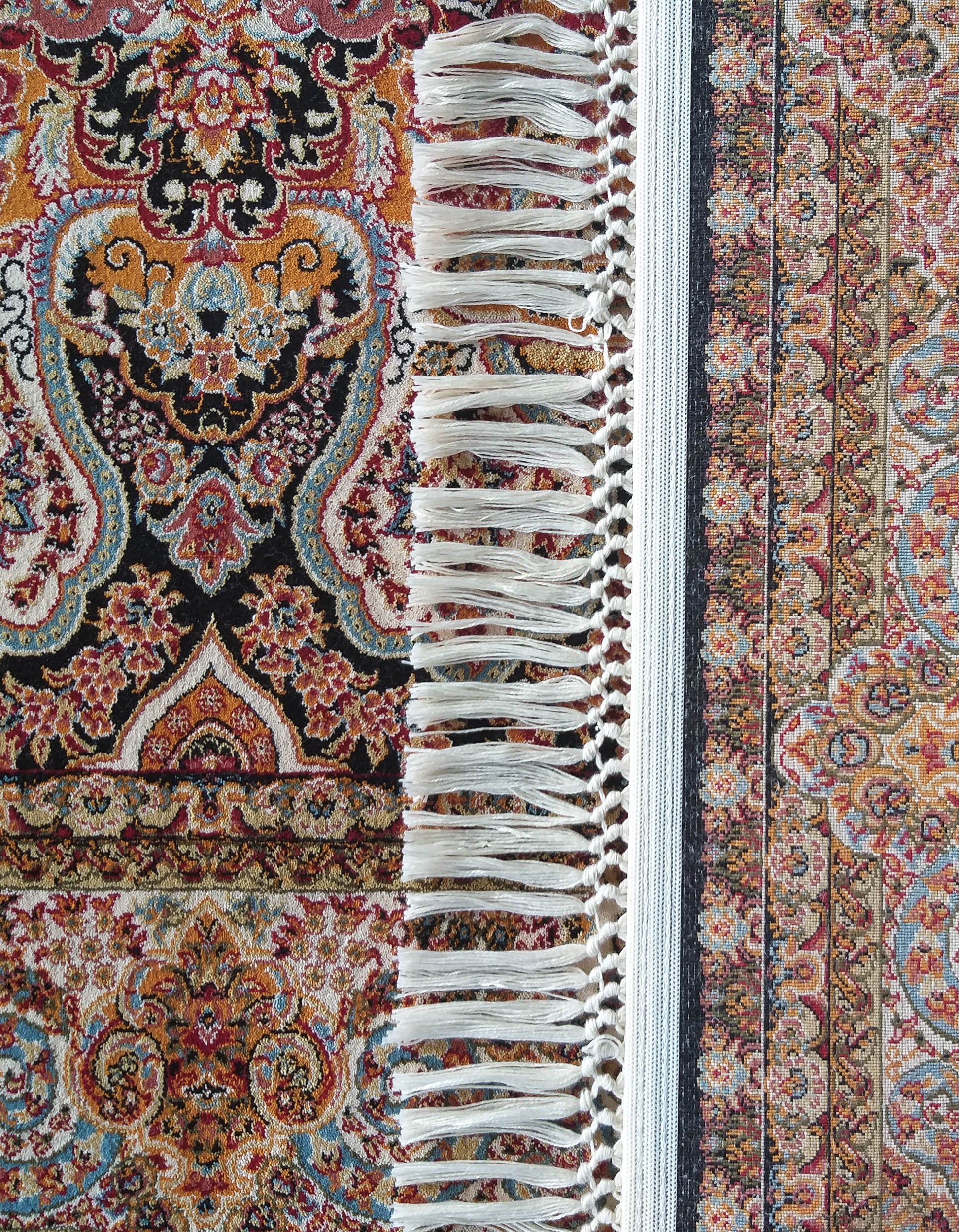 Tapis aux motifs floraux et arabesques entrelacés en rouge, orange, noir, crème et bleu, bordé de franges blanches