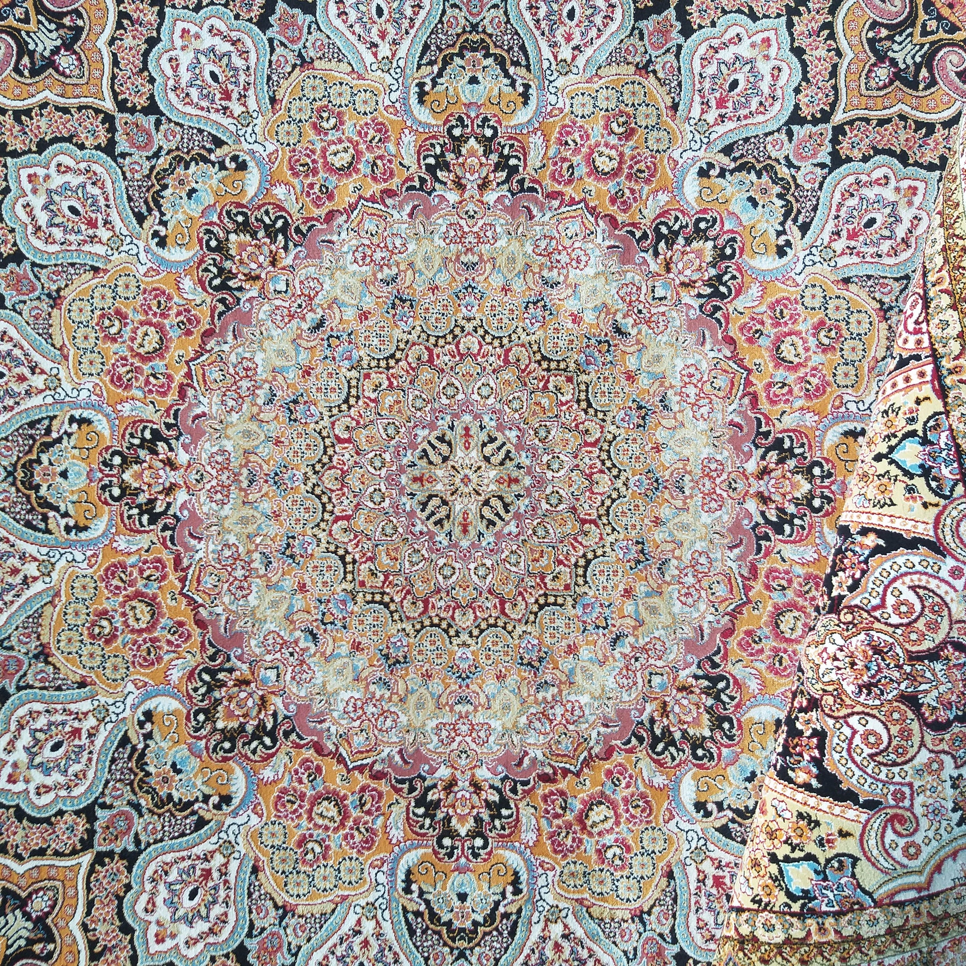Tapis coloré aux motifs orientaux complexes, mêlant rouge, or, noir, bleu et crème. Un bord est replié