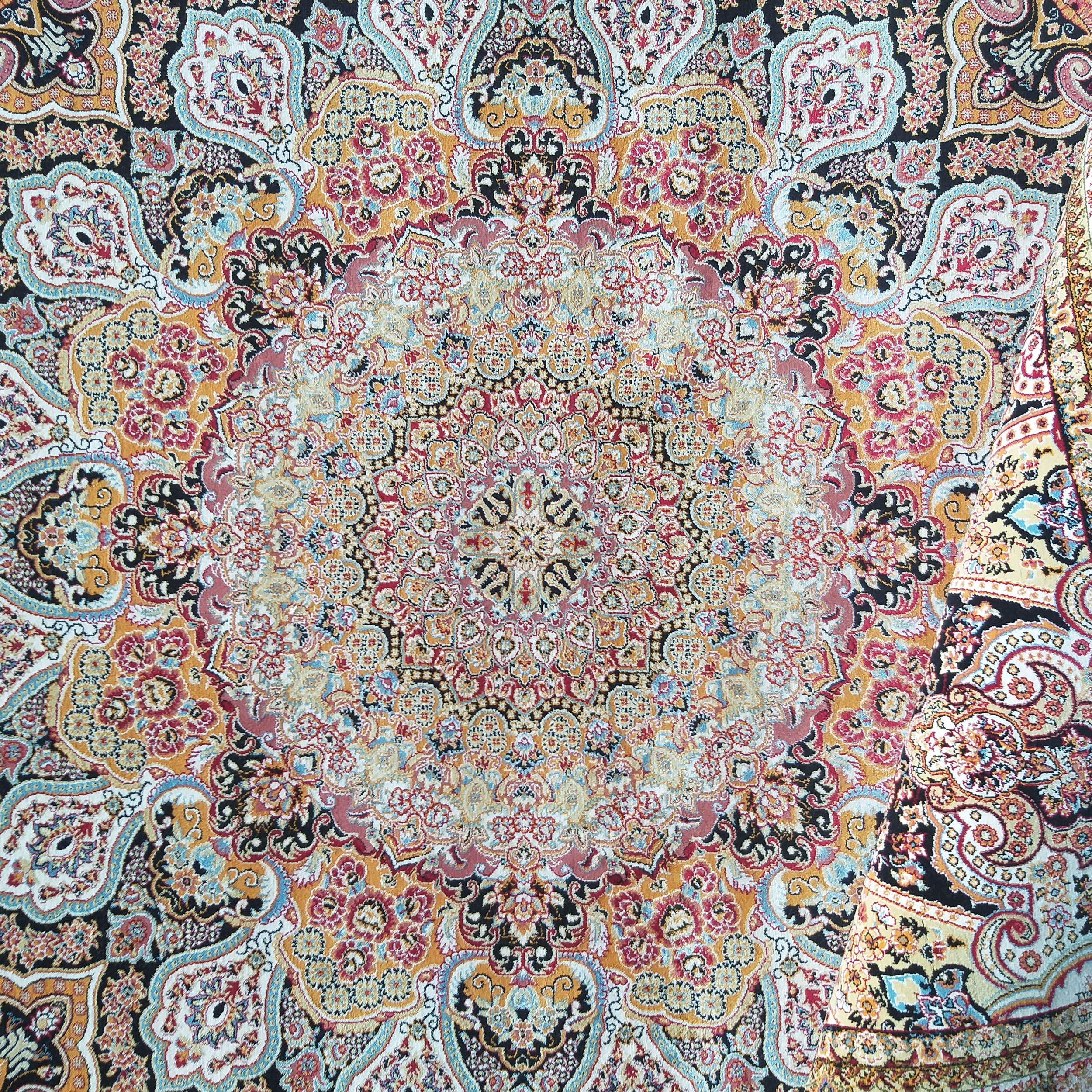 Tapis coloré aux motifs orientaux complexes, mêlant rouge, or, noir, bleu et crème. Un bord est replié