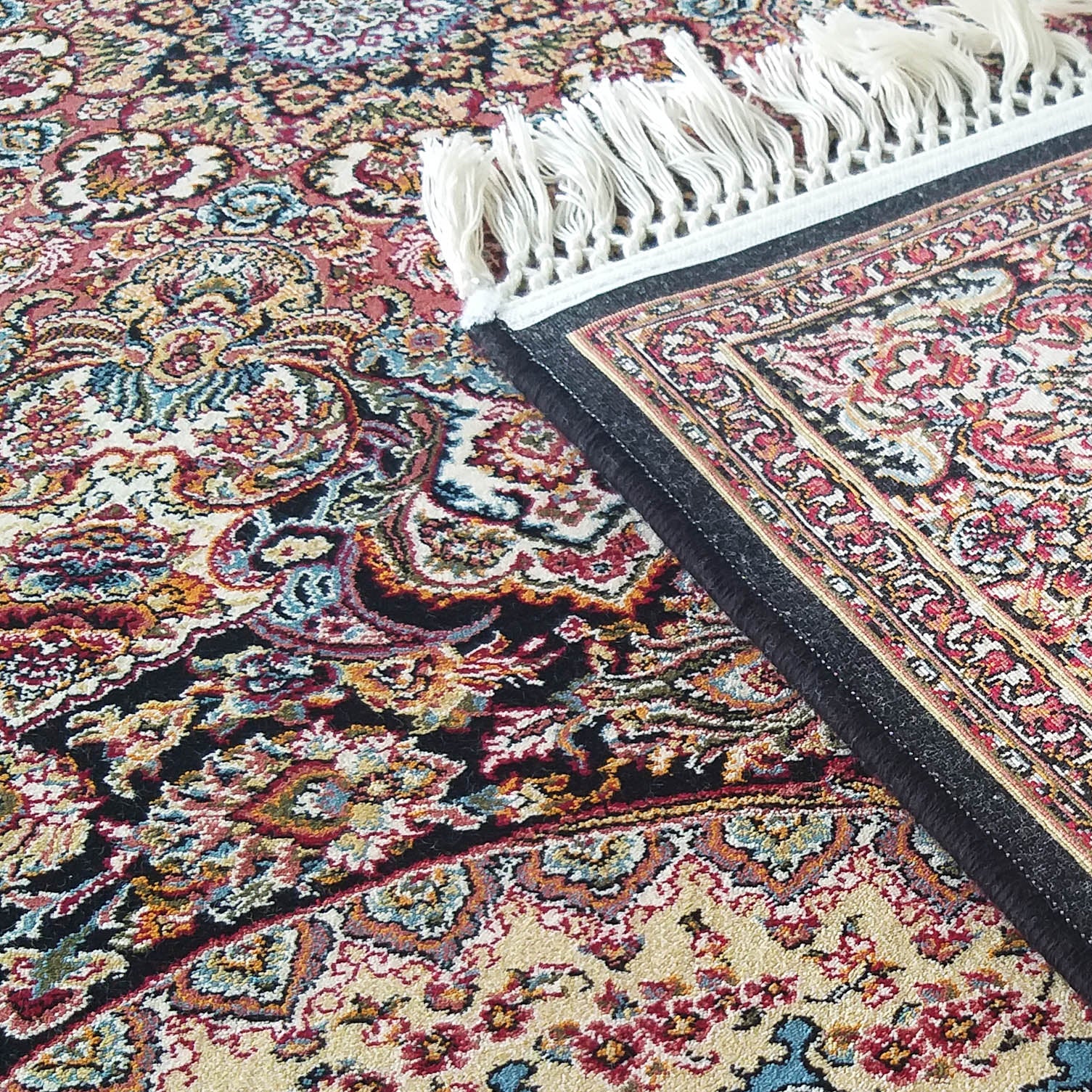 Tapis richement ornementé de motifs floraux complexes et volutes en rouge, bleu, crème, doré et noir, avec des franges