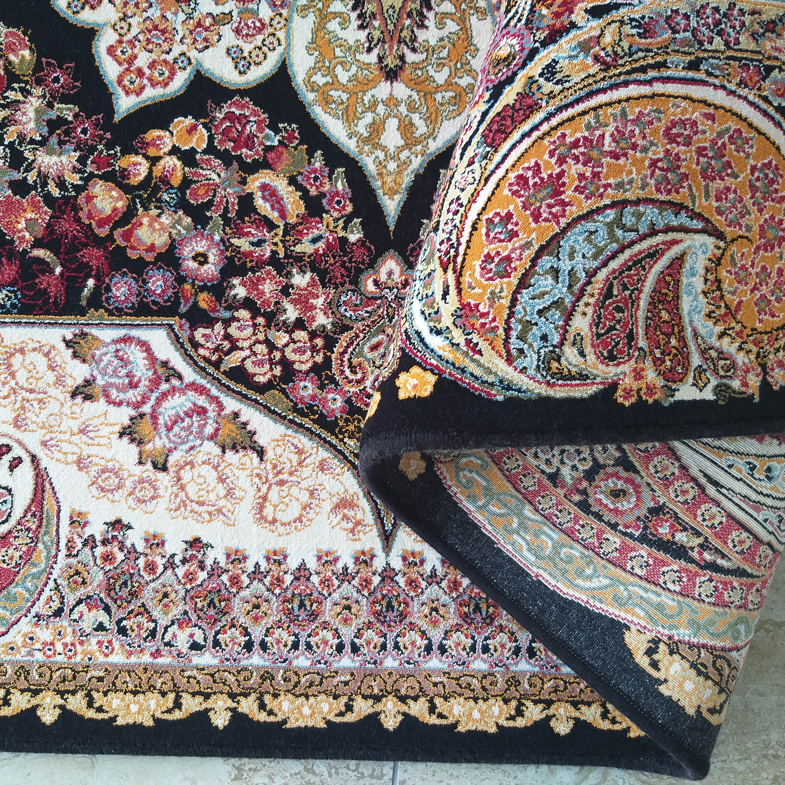Tapis noir à motifs floraux et cachemire rouges, dorés, blancs et bleus. Une partie est roulée, révélant sa surface moelleuse