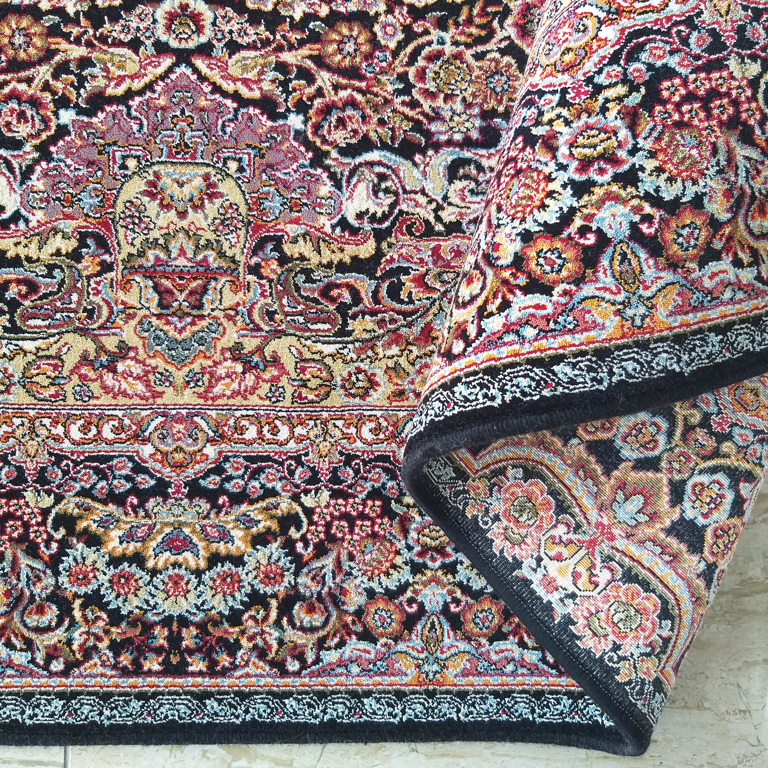 Tapis aux motifs orientaux riches en rouge, or, bleu et blanc sur fond noir. Une partie du tapis est roulée