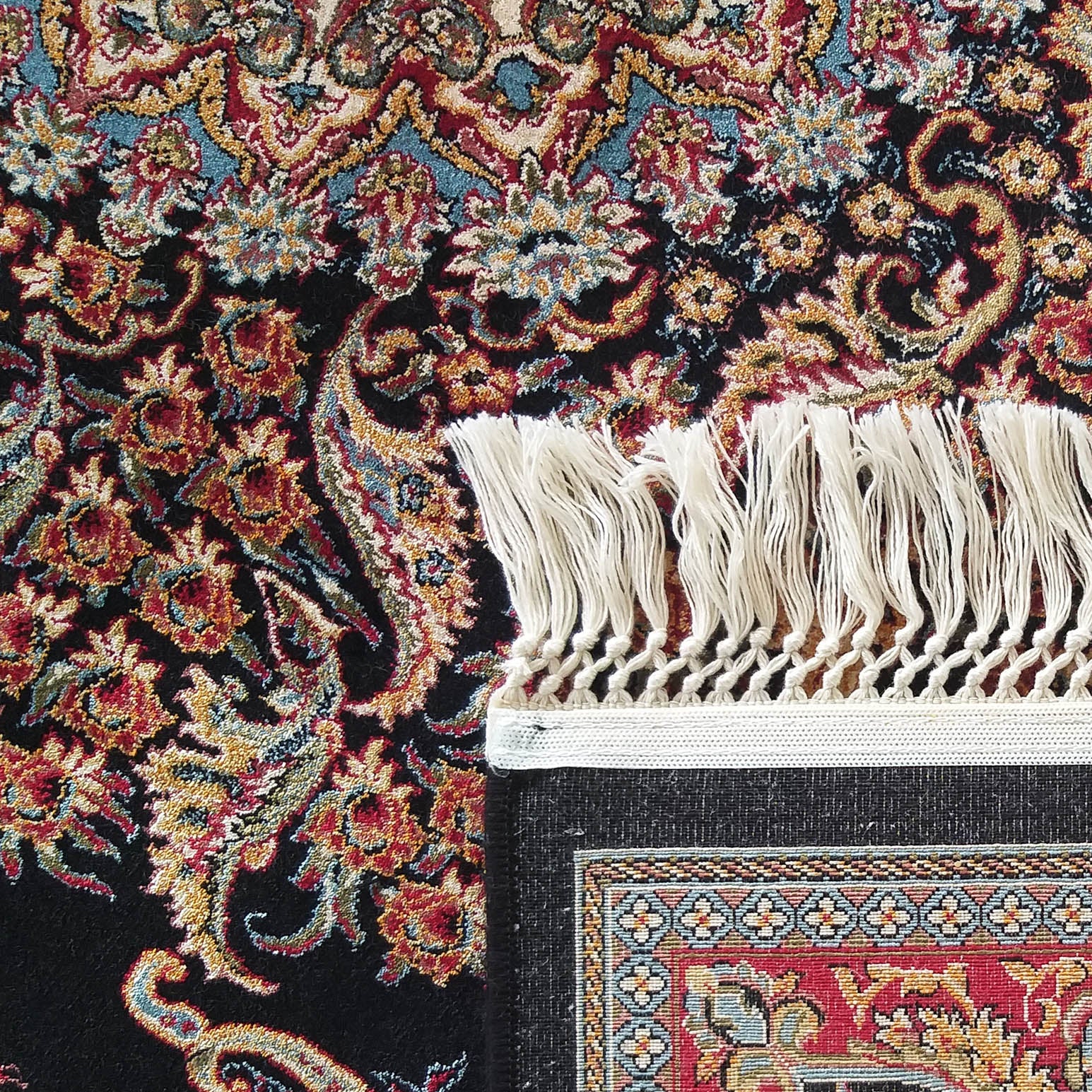 Tapis orné de motifs floraux et arabesques complexes en rouge, or, bleu et vert sur fond noir, bordé de franges claires