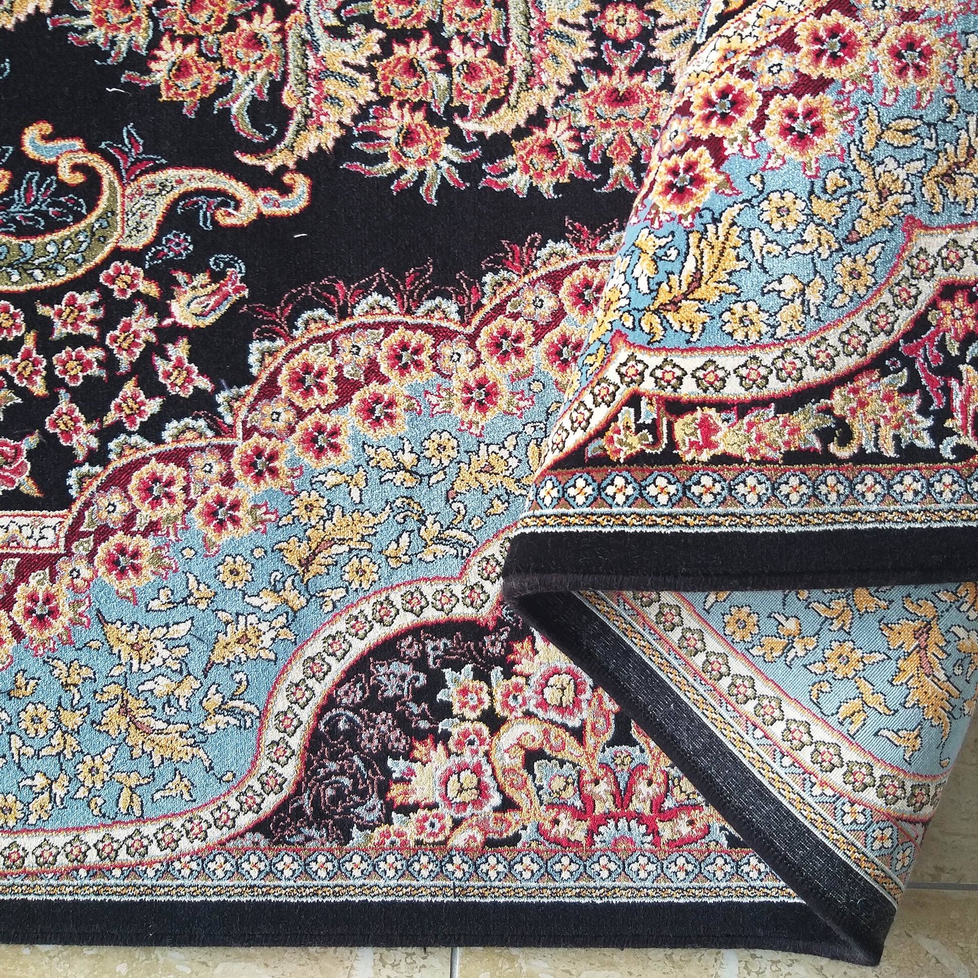 Tapis noir richement décoré de grands motifs floraux et de paisley rouges, dorés et bleus, dont un côté est enroulé