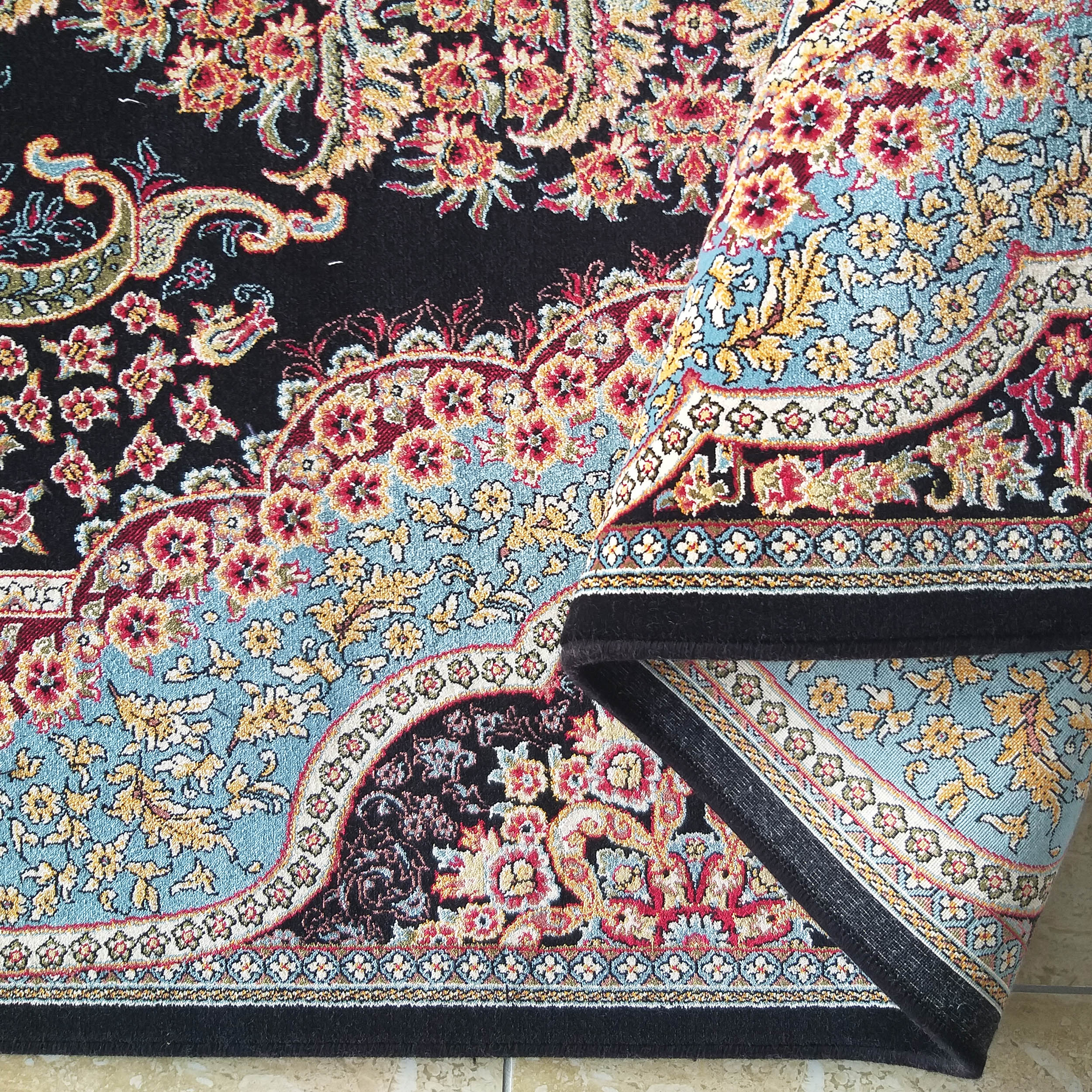 Tapis noir richement décoré de grands motifs floraux et de paisley rouges, dorés et bleus, dont un côté est enroulé