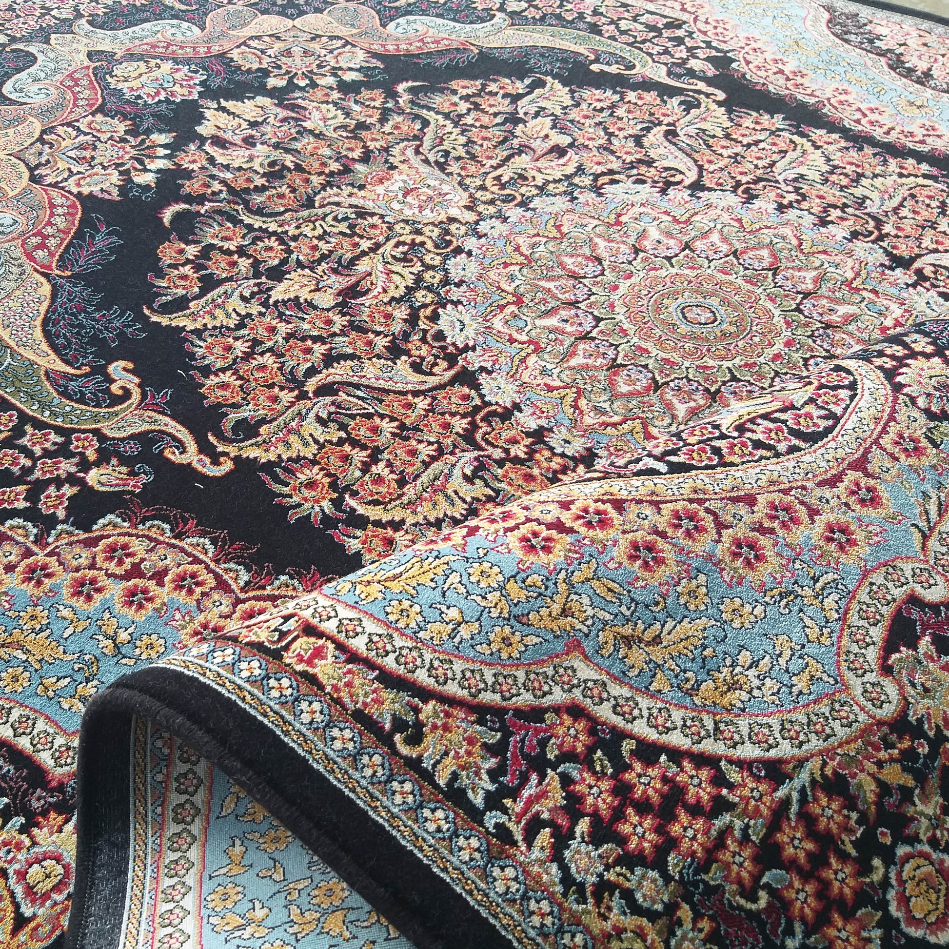 Tapis richement orné de motifs floraux et cachemire rouges, dorés et bleus sur fond noir, une partie est repliée