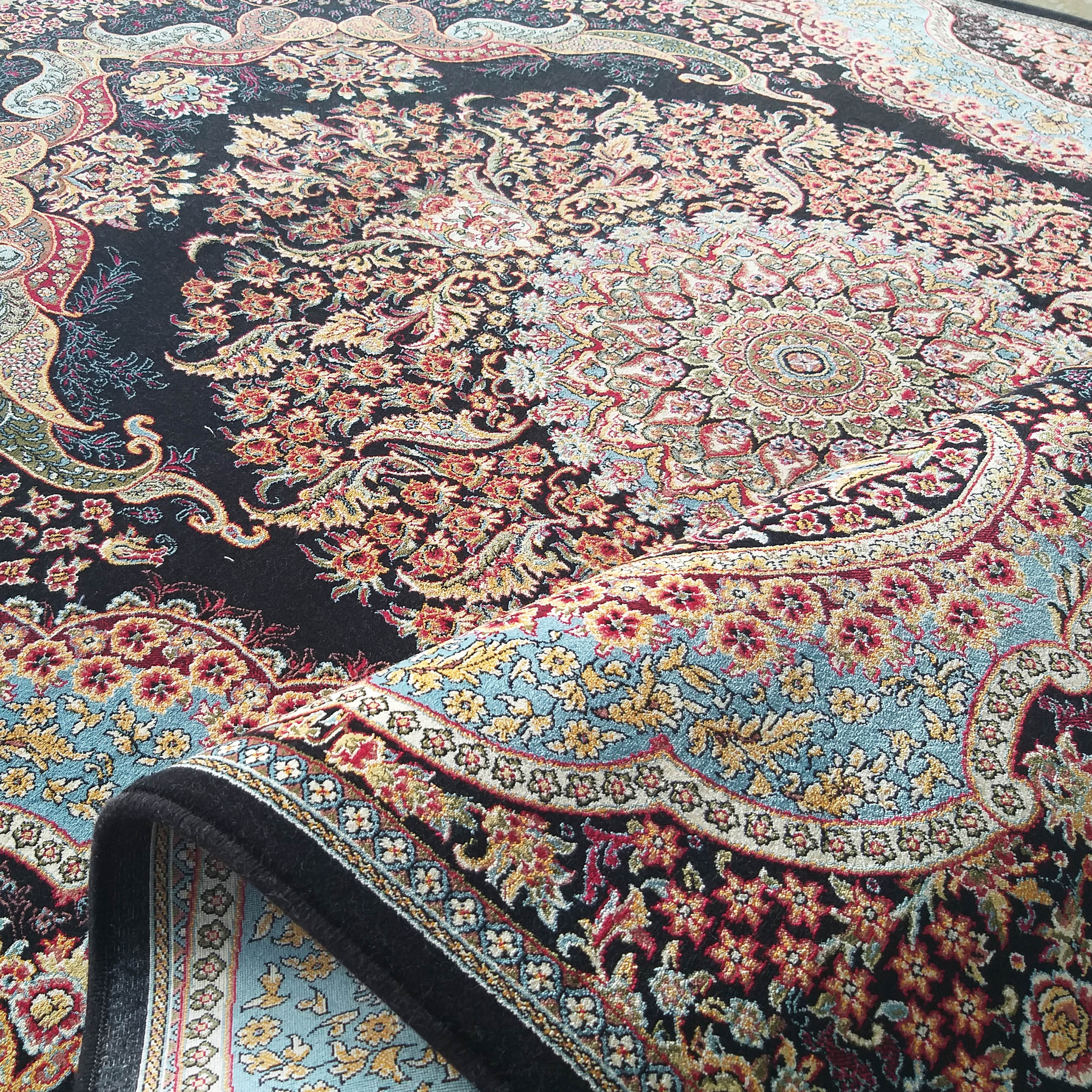 Tapis richement orné de motifs floraux et cachemire rouges, dorés et bleus sur fond noir, une partie est repliée