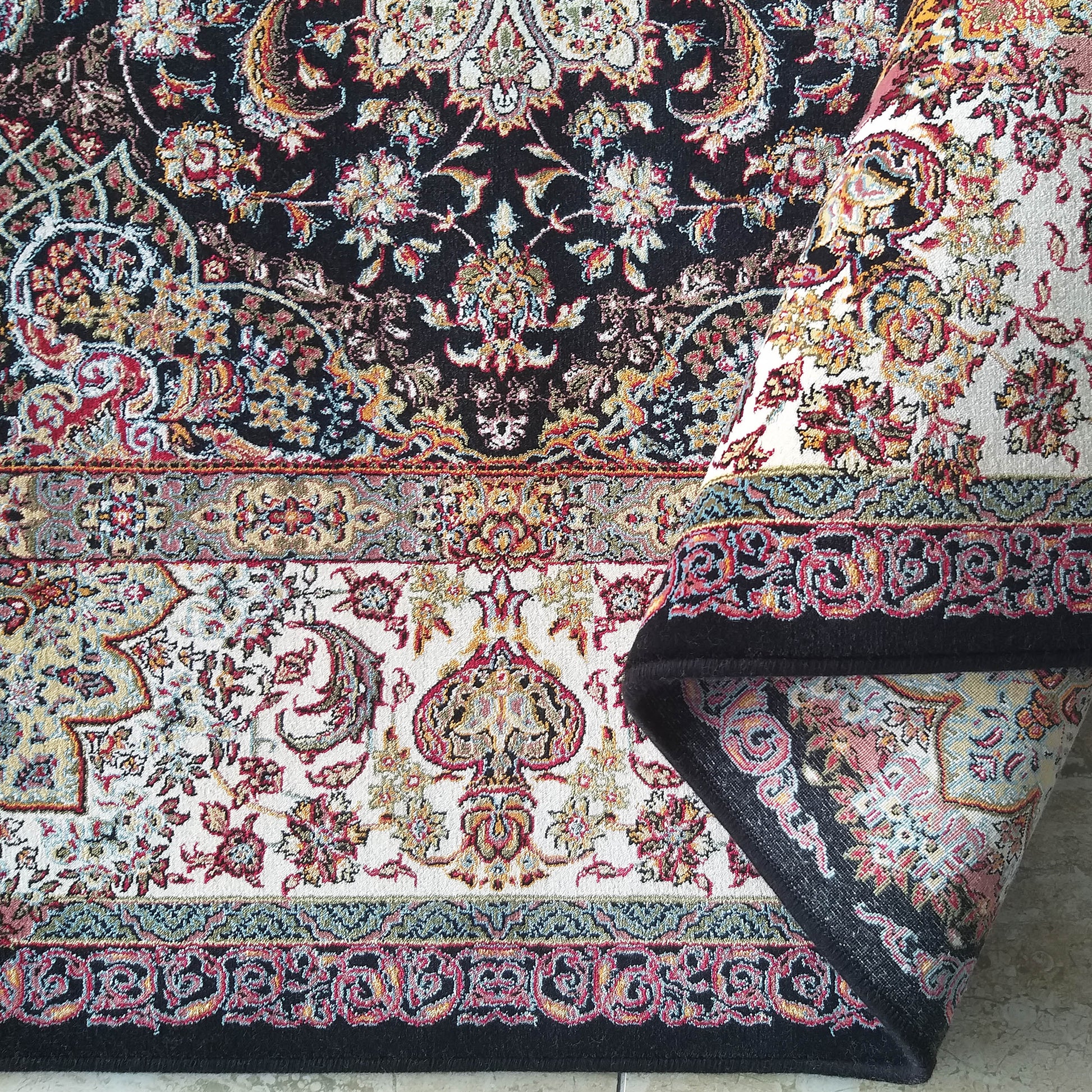 Tapis à motifs floraux et cachemire sur fonds noir et blanc crème. Des couleurs rouge, or et bleu ressortent. Une partie du tapis est roulée