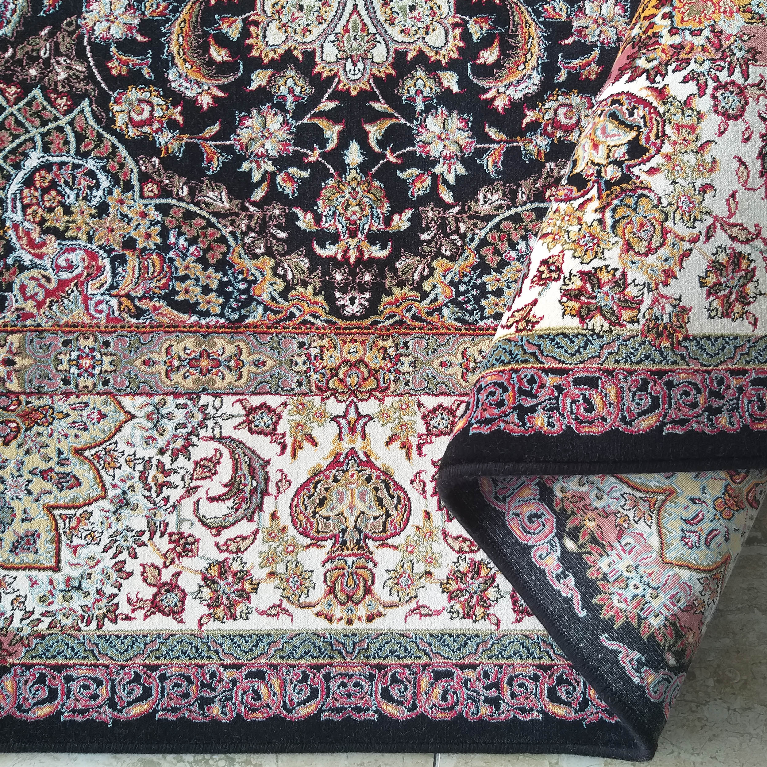 Tapis à motifs floraux et cachemire sur fonds noir et blanc crème. Des couleurs rouge, or et bleu ressortent. Une partie du tapis est roulée