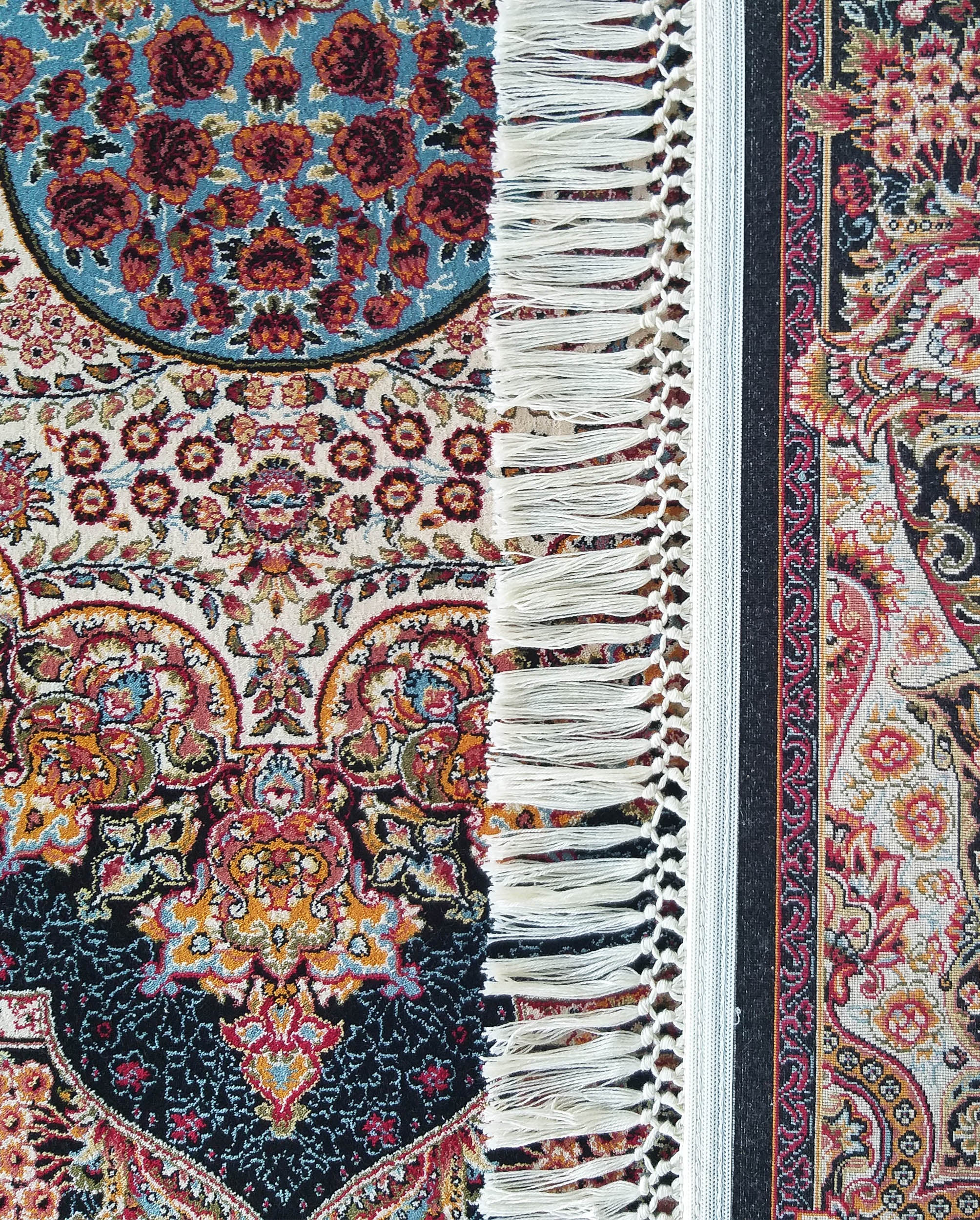 Tapis richement orné de motifs floraux complexes en rouge, or, bleu et crème sur fonds noir et blanc. Des franges claires bordent la pièce