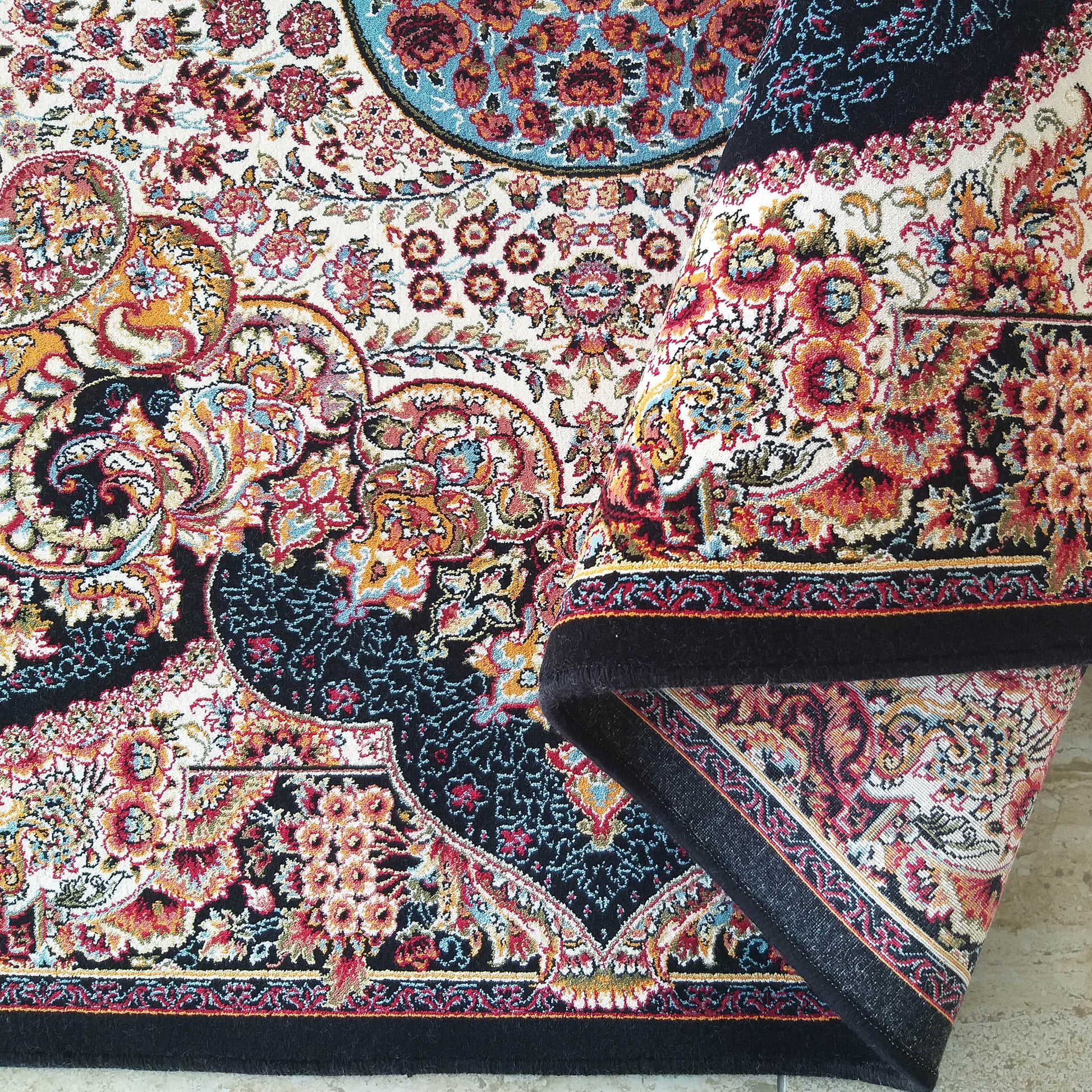 Tapis coloré à motifs floraux et volutes, en noir, crème, rouge, orange et bleu. Une partie du tapis est roulée